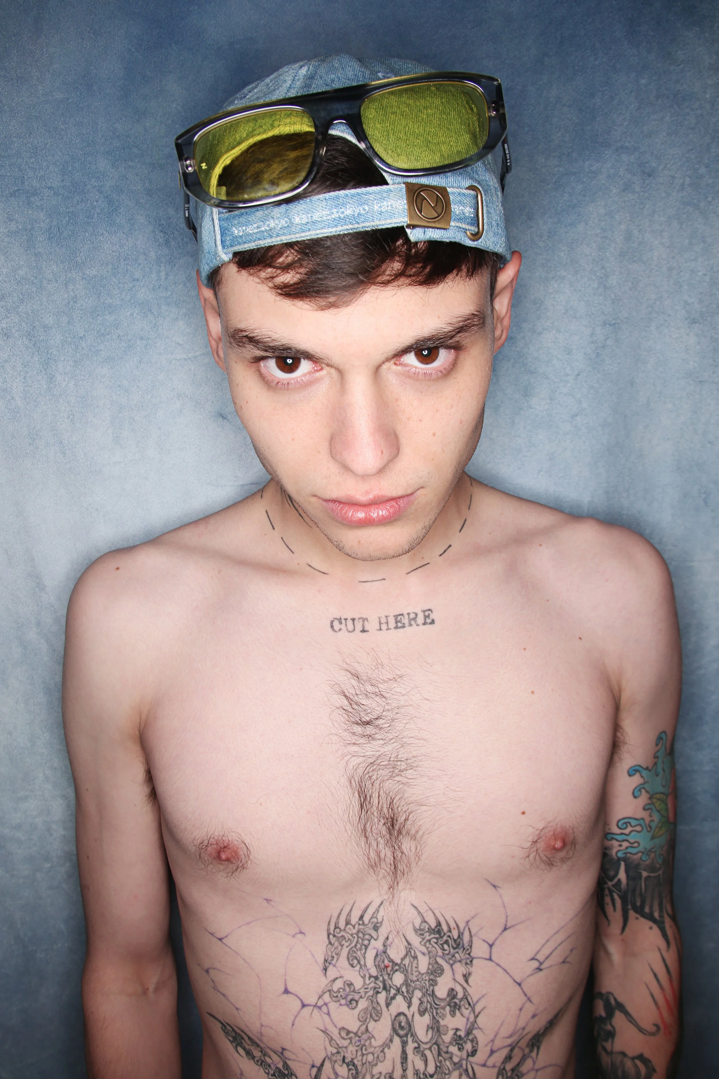 A shirtless man lying against a blue background, wearing sunglasses with yellow lenses and a denim cap. He has tattoos on his chest and arms, including a phrase 'CUT HERE' on his chest and intricate designs on his torso and right arm.