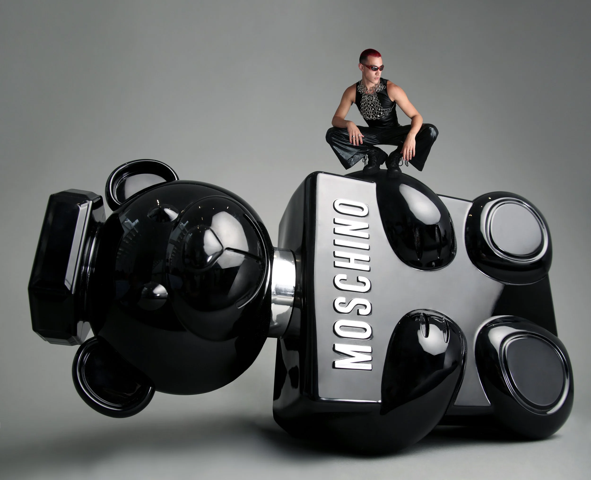 Close-up of a glossy black and white car sculpture with a person squatting on top, wearing sunglasses, black leather clothing, and a metal chain necklace, against a plain gray background.