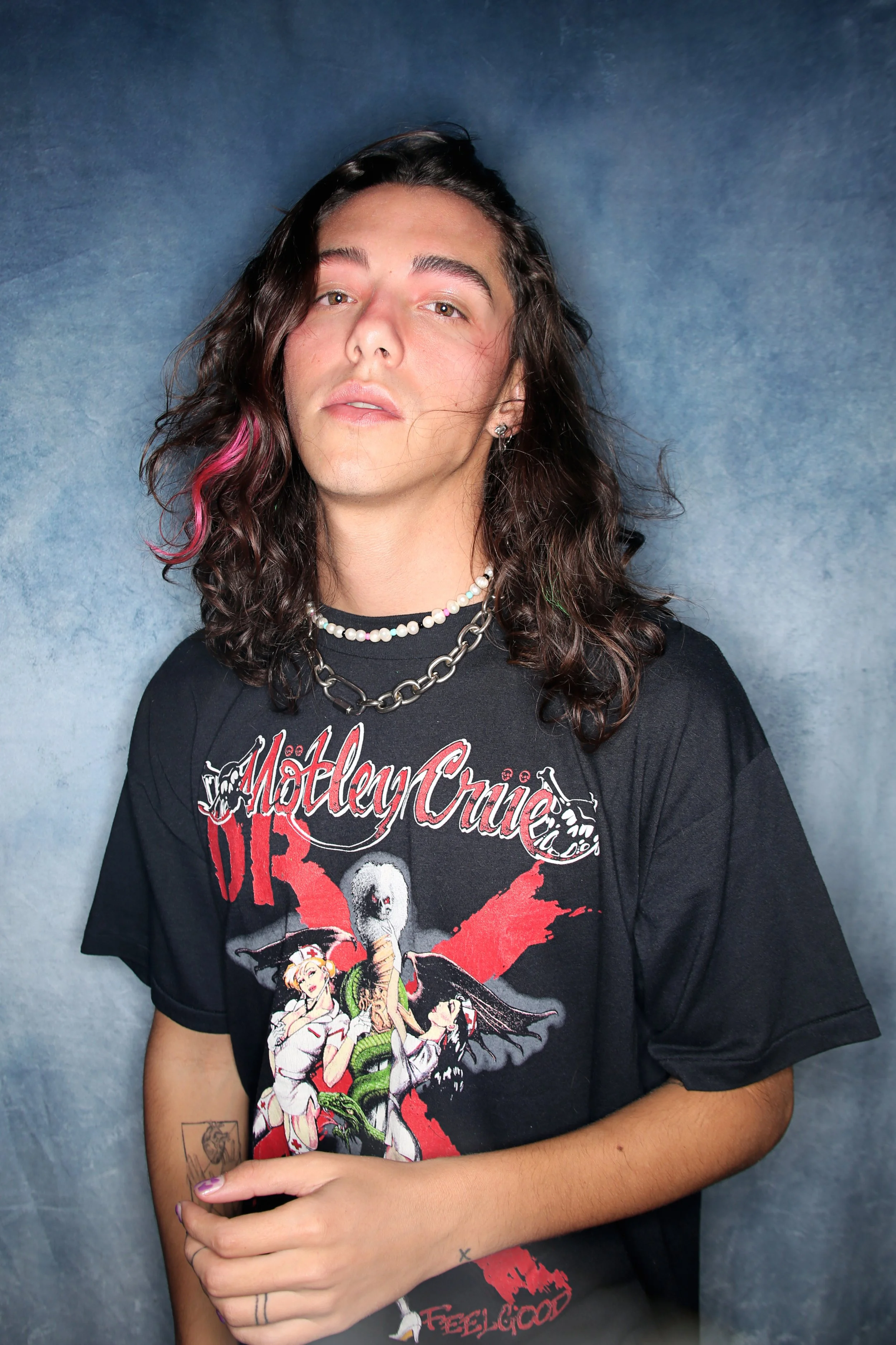 A person with long, wavy dark hair accented with hints of pink, wearing a black Motley Crue band t-shirt, layered chains, a pearl necklace, and earrings, standing against a textured blue background.