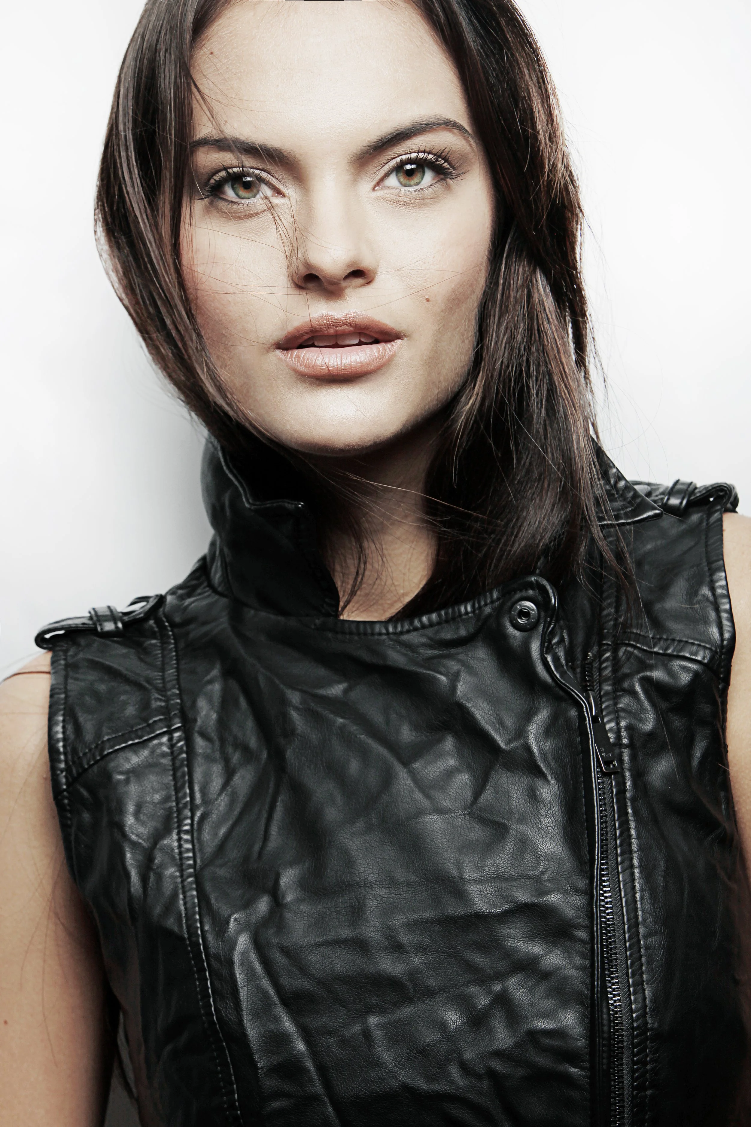 Close-up of a woman with brown hair and green eyes wearing a black leather vest against a plain white background.