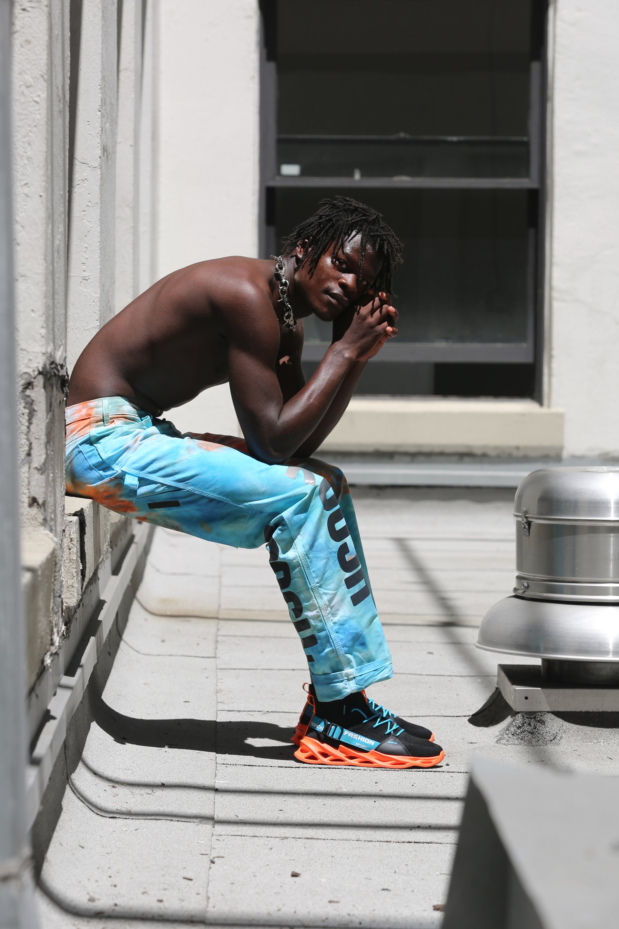 A young man with dreadlocks, shirtless, wearing tie-dye pants, and brightly colored sneakers, sitting on a ledge on a rooftop or terrace, looking towards the camera with hands clasped.