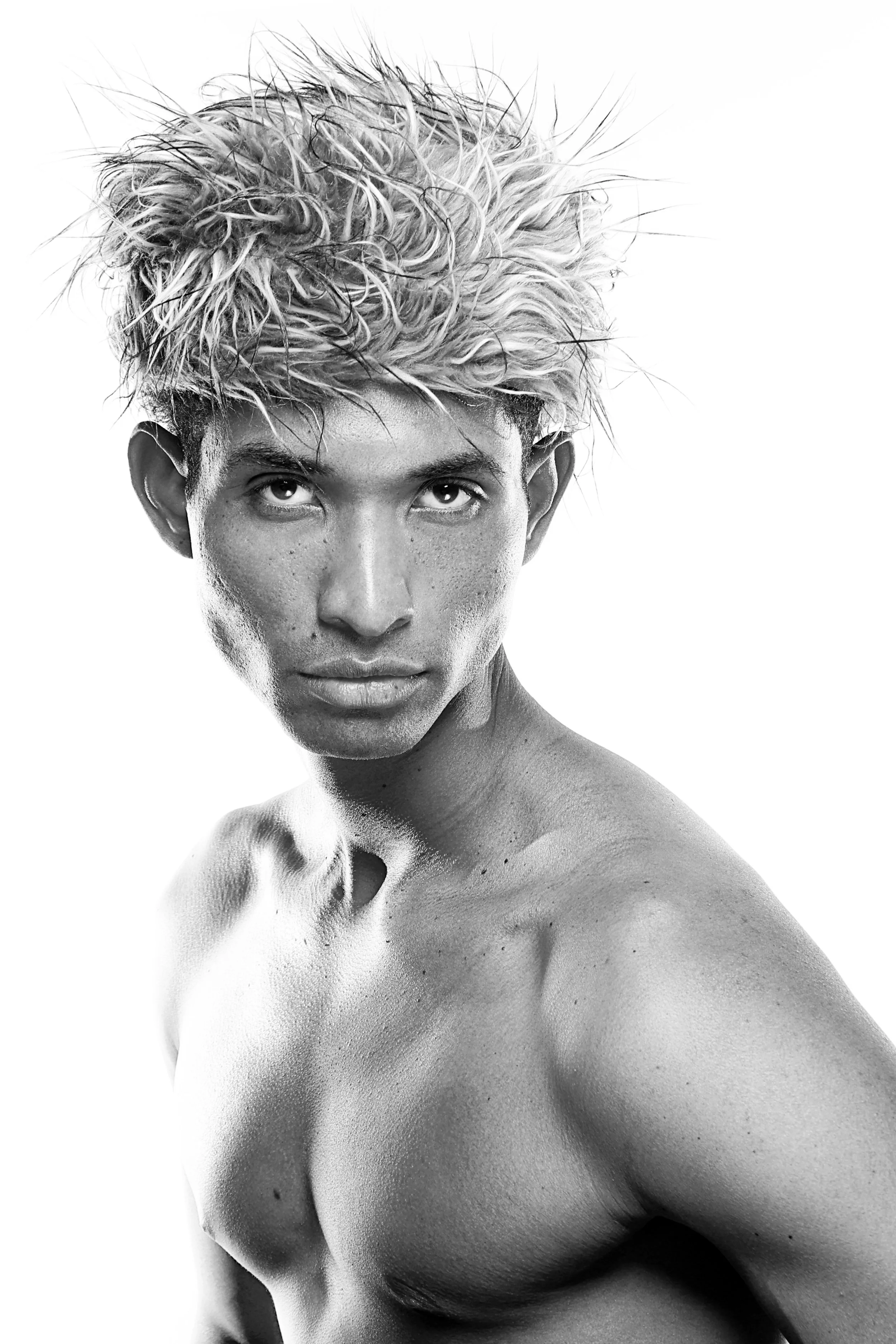 Black and white photo of a shirtless man with tousled, spiky hair, looking directly at the camera with intense gaze.