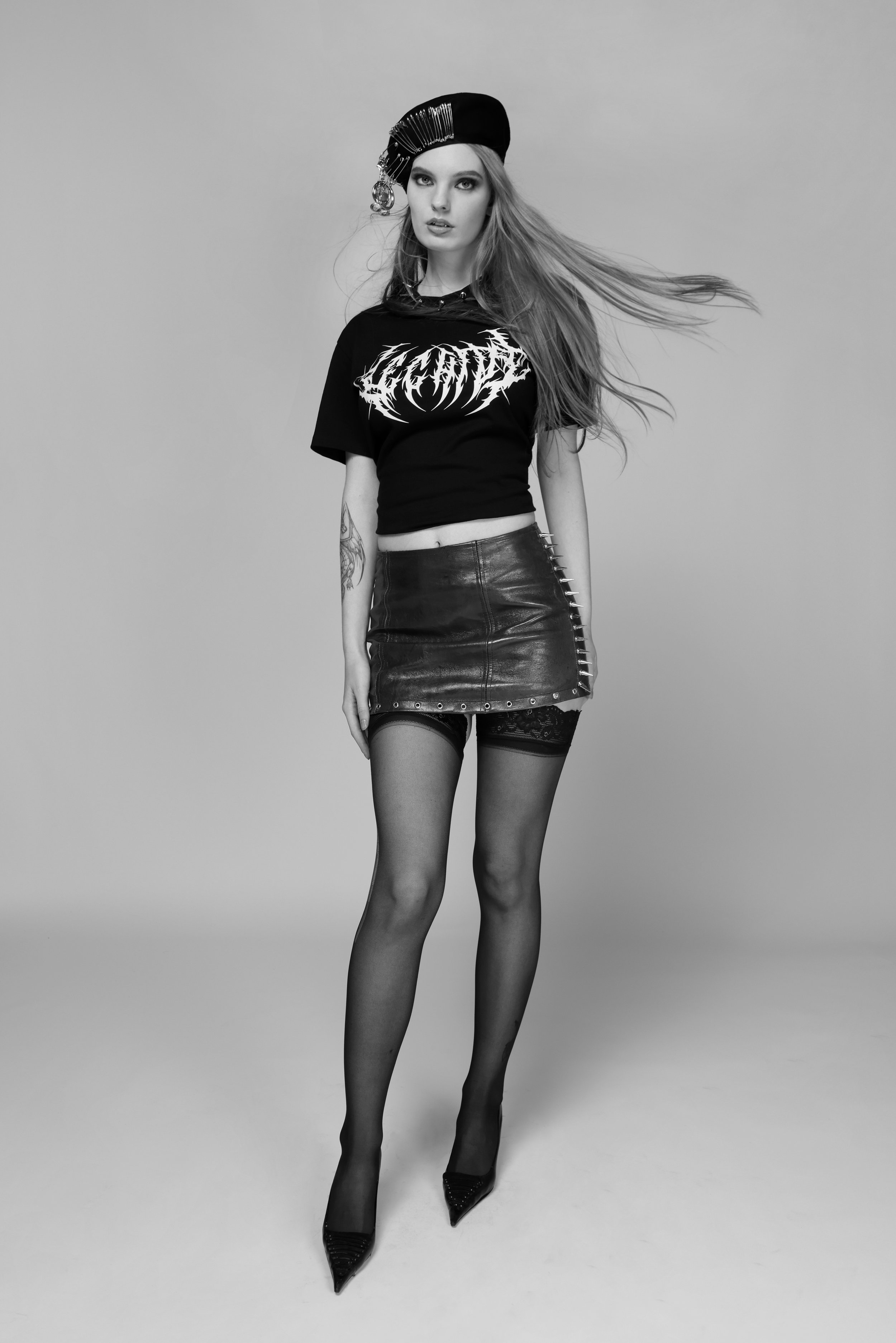 Black and white photograph of a young woman with long hair, wearing a black beret with decorative pins, a T-shirt with an intricate logo, a leather skirt with metal spikes on the side, fishnet stockings, and high heels, standing against a plain backg