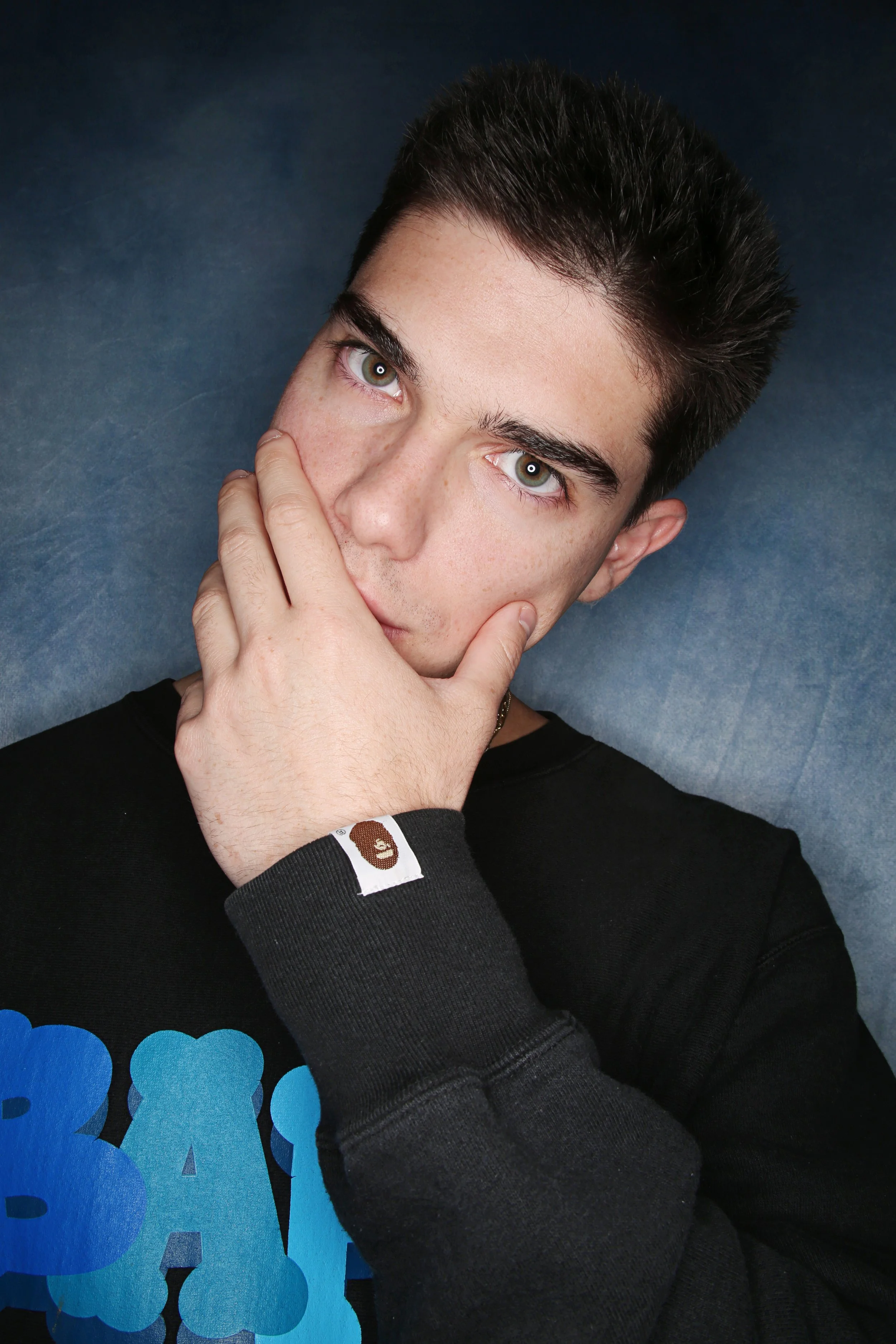 A young man with dark hair and light skin is posing with his hand covering part of his face, looking directly at the camera. He is wearing a black shirt with blue designs and a black wristband with a brown bear logo. The background is a dark blue tex
