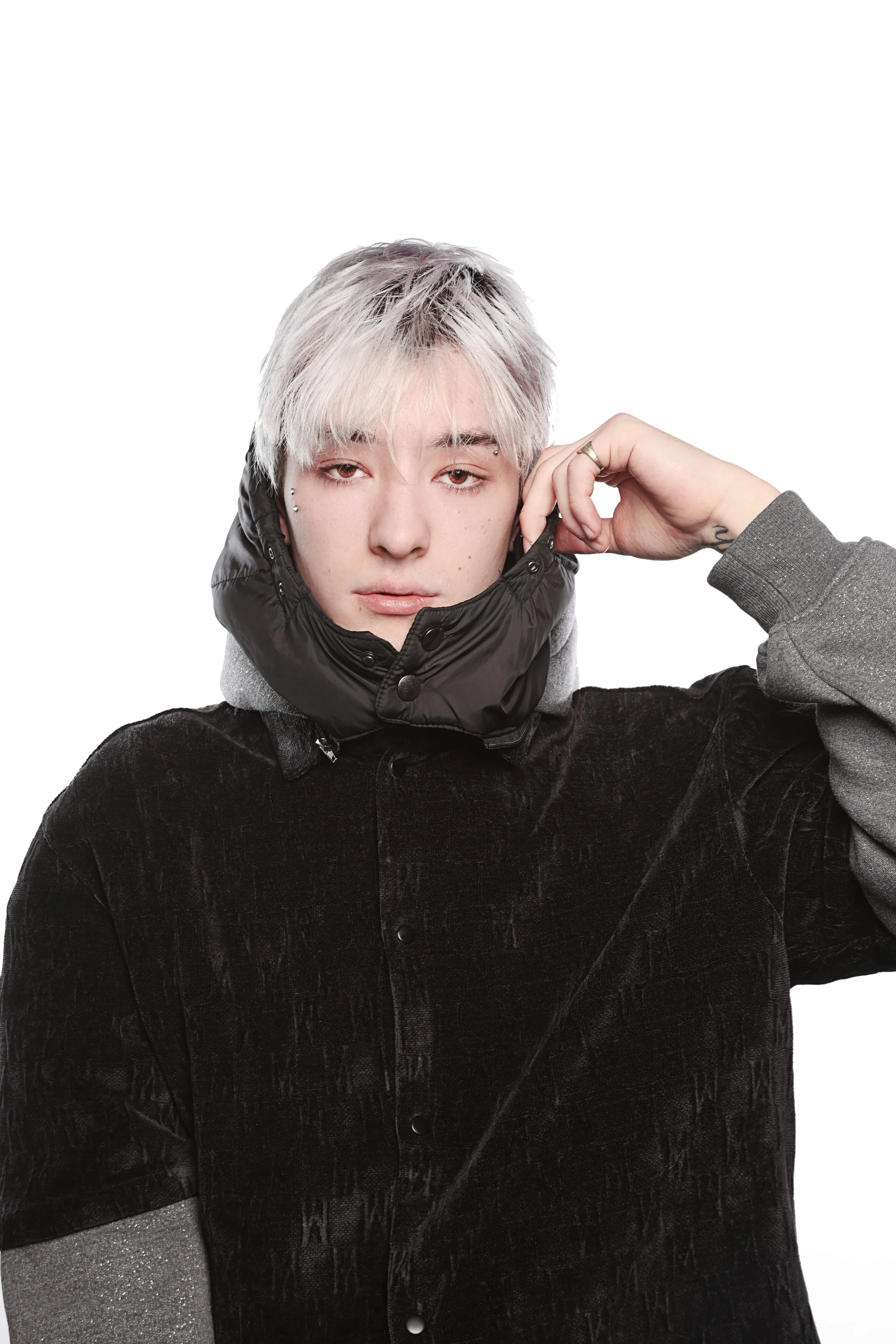 A person with platinum blonde hair wearing a black puffer jacket and a gray hoodie, adjusting the hood with one hand against a plain white background.