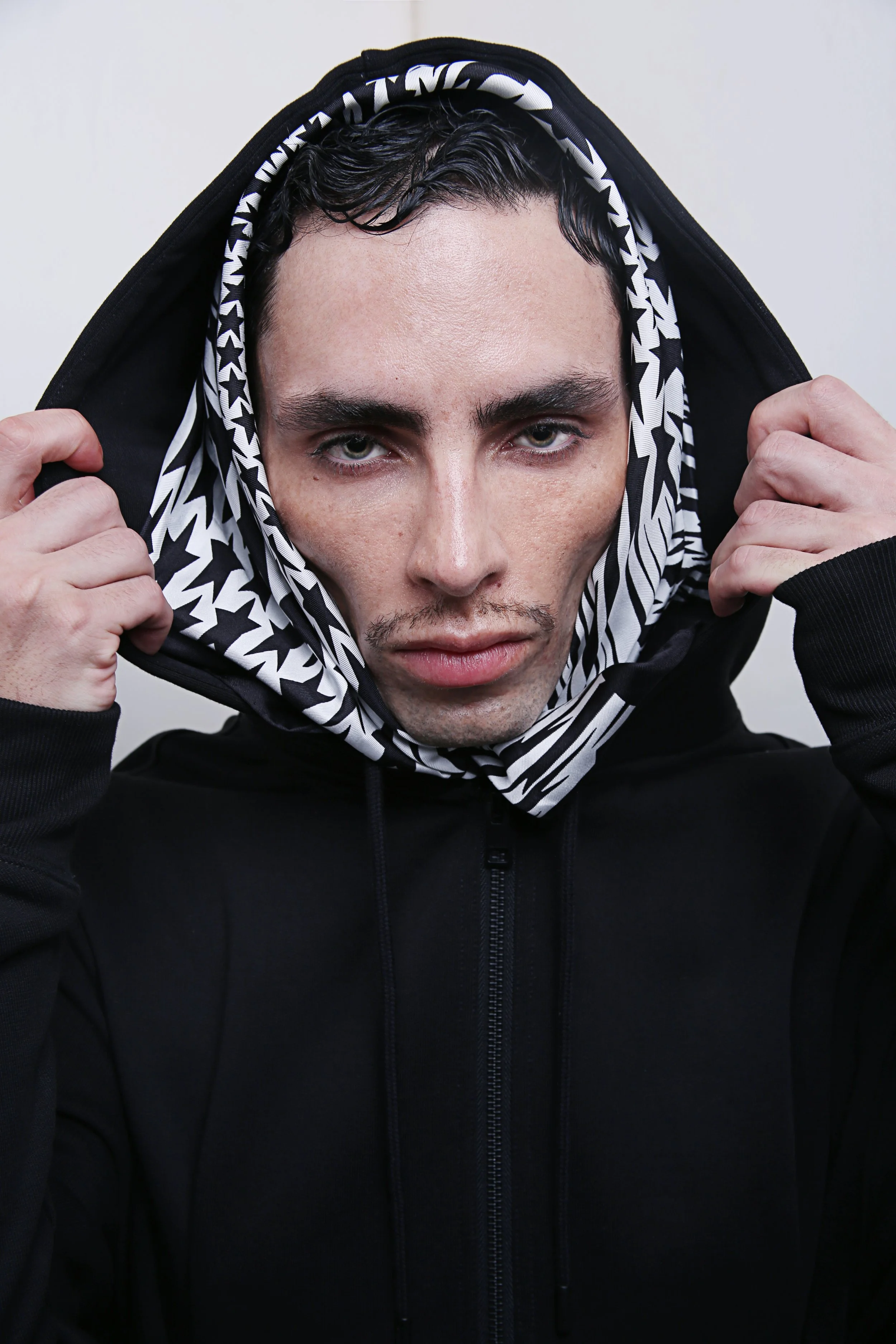 A young man with dark, wet, curly hair wearing a black hoodie with a patterned lining in black and white, holding the hood with both hands, in front of a plain white background.