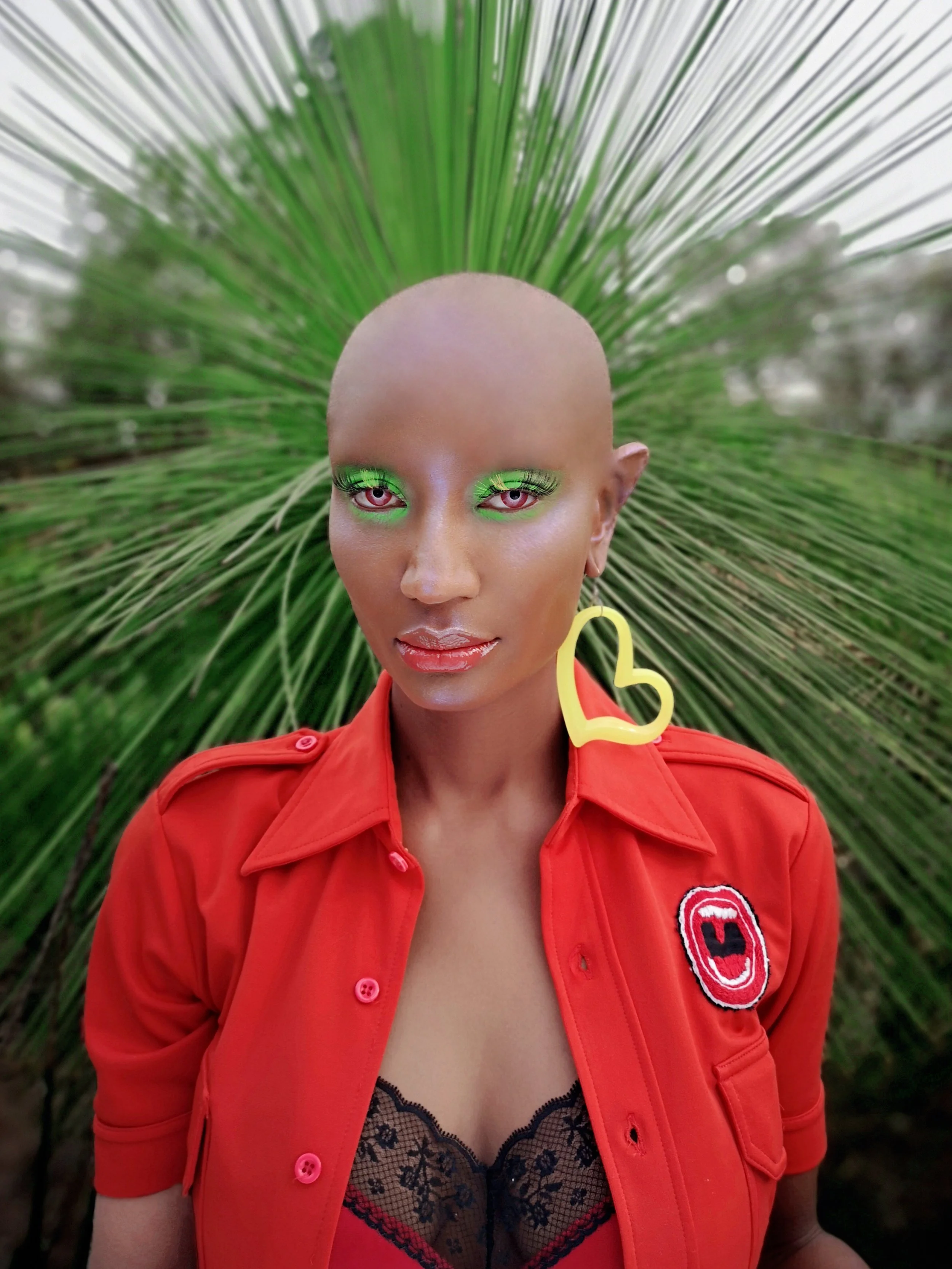 A person with light skin wearing neon green eyeshadow, pink eyes, and a neon yellow heart-shaped earring, dressed in a red shirt with a patch on the sleeve, standing in front of green palm leaves.
