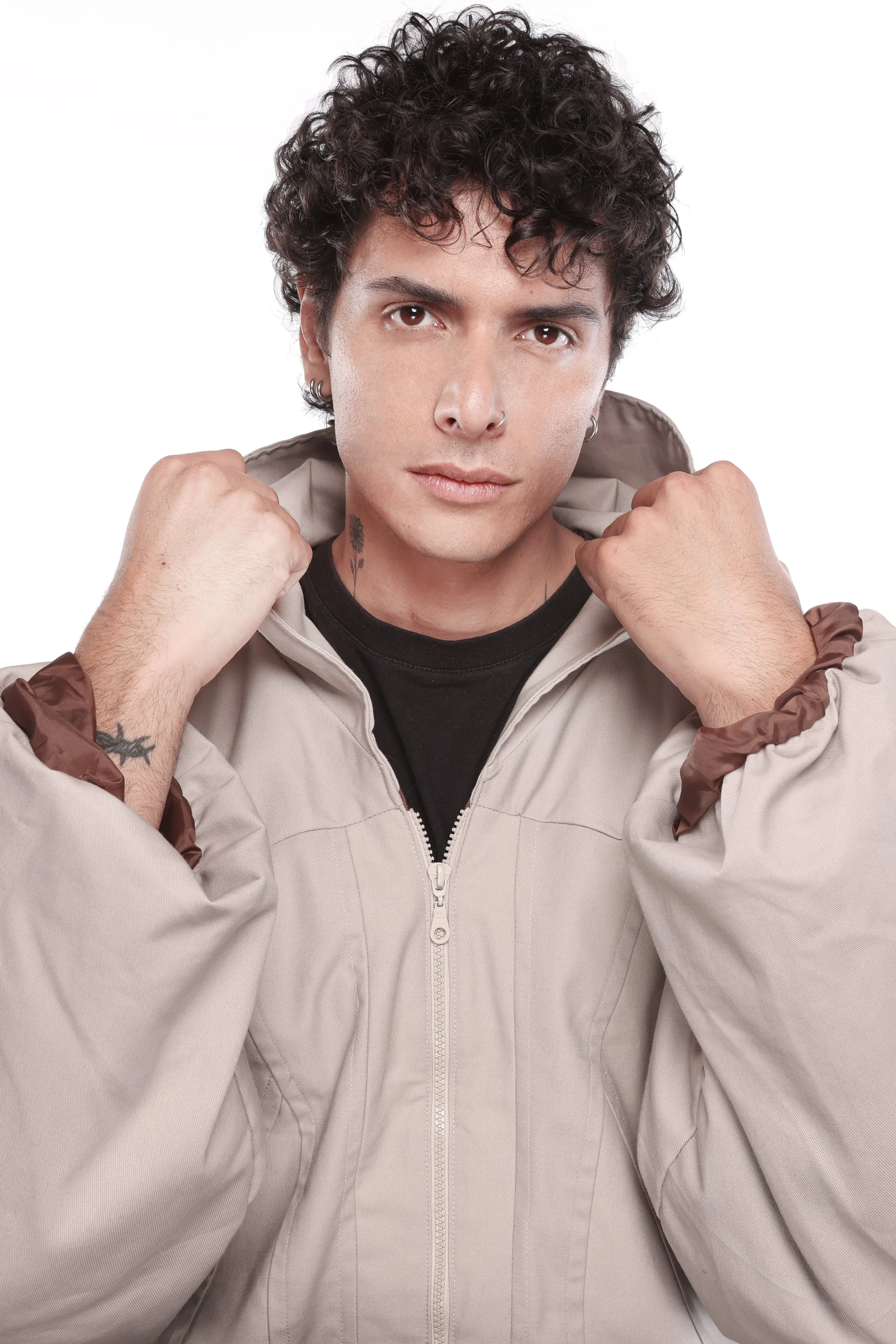 A young man with curly dark hair, light skin, and multiple ear piercings, wearing a beige zip-up jacket with brown accents, holding the collar of the jacket with both hands. He is looking directly at the camera against a white background.
