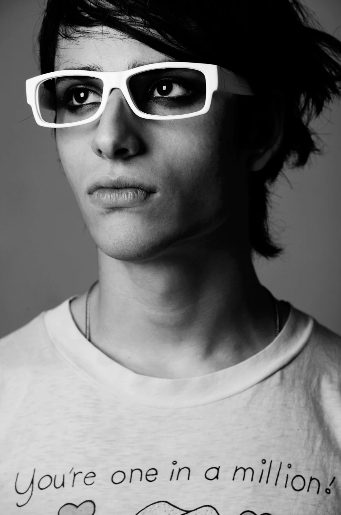 A person with short dark hair wearing white glasses looking slightly upward. They are wearing a light-colored shirt with text and a heart graphic on it.