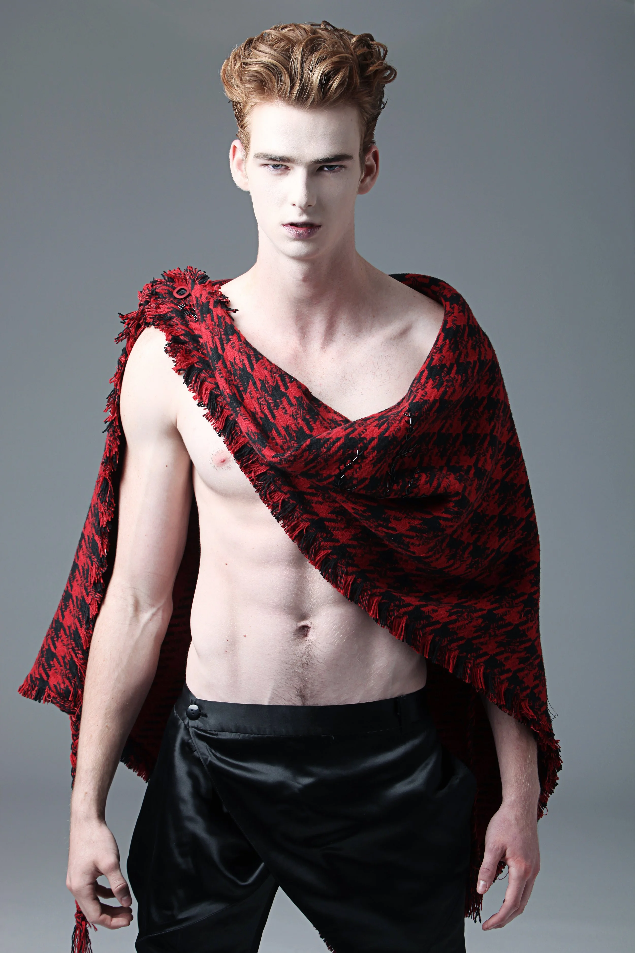 A young man with styled blonde hair, wearing a red and black patterned shawl draped over one shoulder, exposing his bare chest, and black shiny pants, against a plain gray background.
