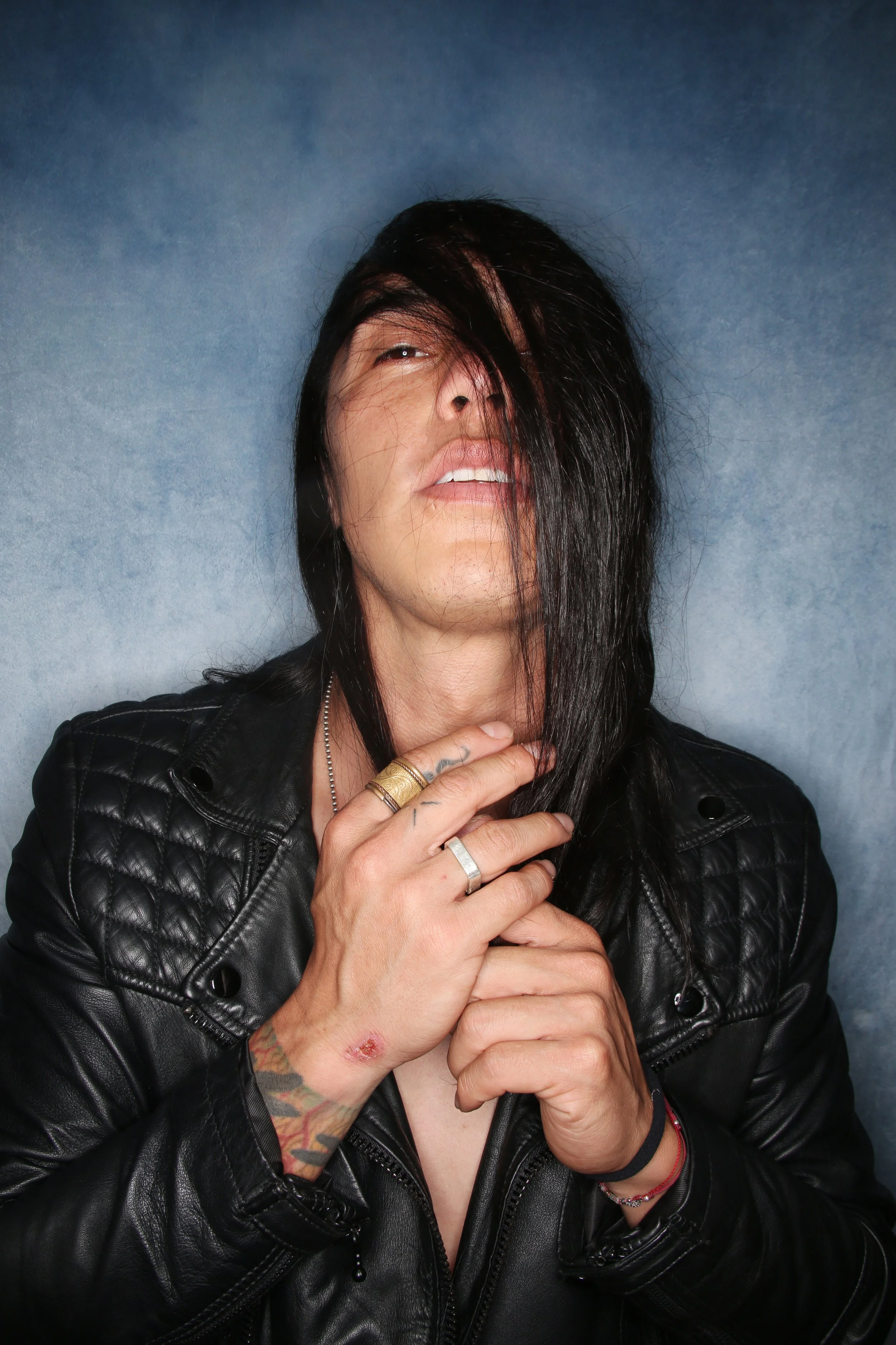 A woman with dark hair and tattoos on her hands and arm, wearing a black leather jacket and multiple rings, striking a pose with her hands near her face and hair partially covering her face against a grey background.