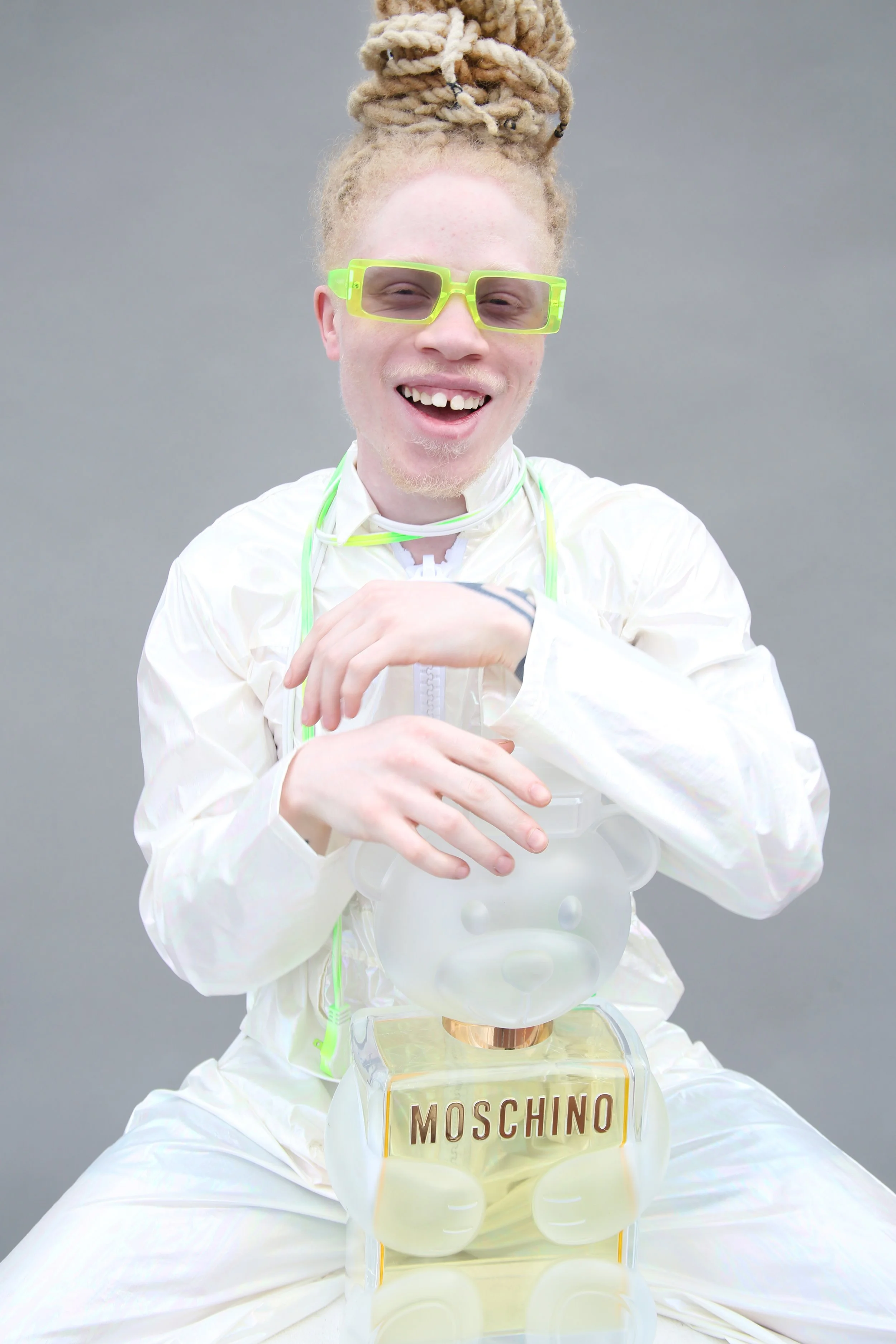 Person with blond dreadlocks wearing yellow-green glasses and a cream-colored outfit, smiling and sitting with a Moschino perfume bottle in front of them, against a light gray background.