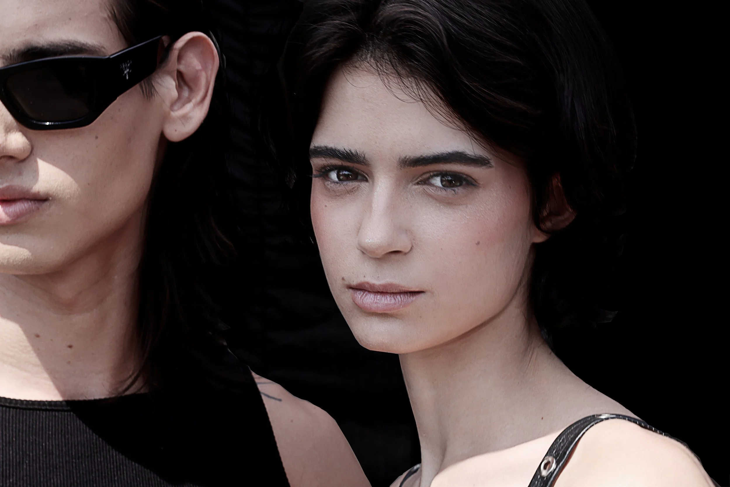 Close-up of two women with dark hair and fair skin, one wearing sunglasses, against a black background.