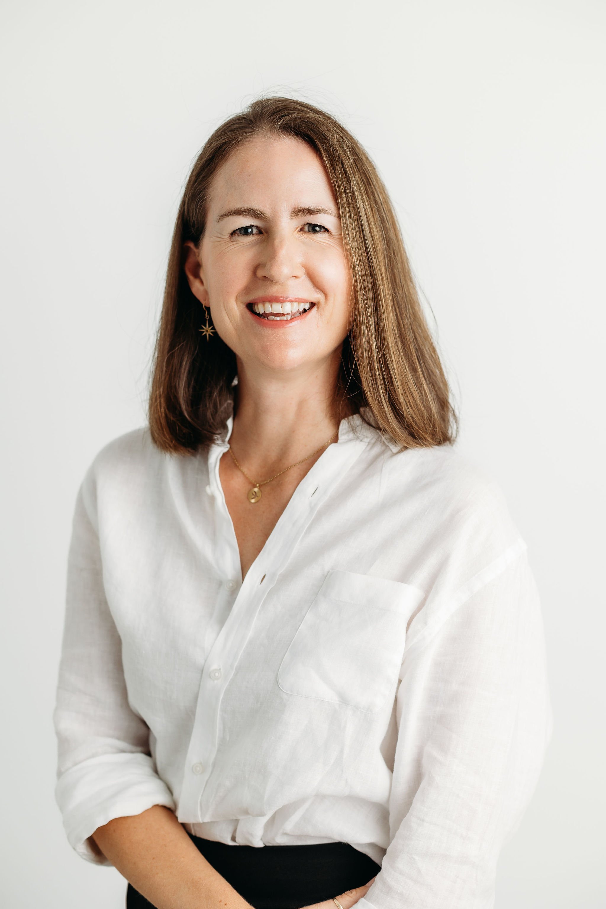 A woman with shoulder-length brown hair, wearing a white button-up shirt, smiling and looking at the camera, with a plain white background.