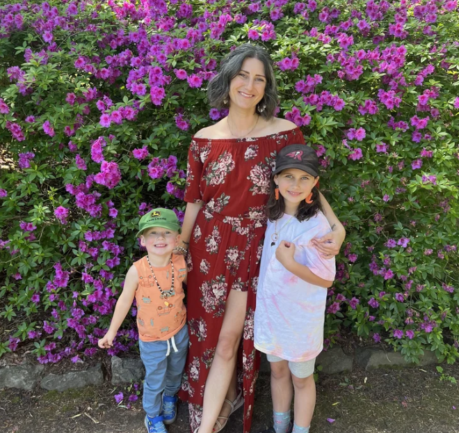 A woman and two young girls standing in front of pink flowers. The woman is wearing a red floral dress and has shoulder-length dark hair, smiling. The girl on the left is wearing an orange tank top, blue pants, and a green hat, smiling. The girl on the right is wearing a white T-shirt, light-colored shorts, and a black cap, also smiling.