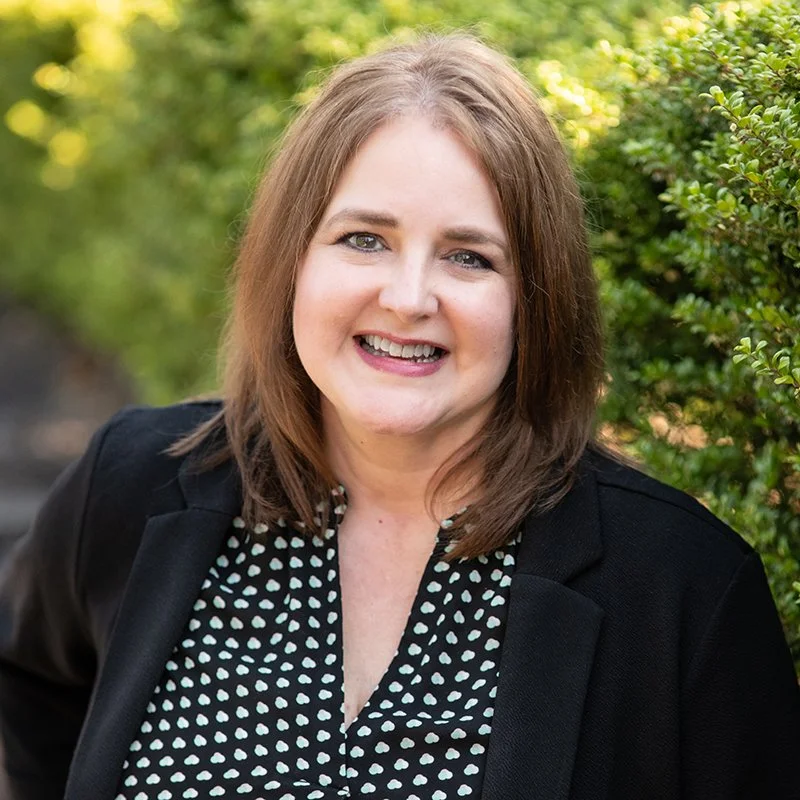 A woman with shoulder-length brown hair smiling, wearing a black blazer over a black and white polka dot blouse, standing outdoors near green bushes.