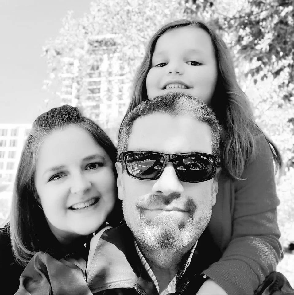 A family of three, a man, a woman, and a young girl, smiling and posing for a selfie outdoors with trees and a building in the background. The man is wearing sunglasses, the woman has shoulder-length hair, and the girl has long hair.