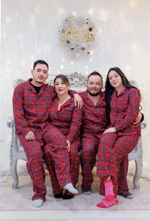 Four people in matching red plaid pajamas sitting on a vintage white and silver sofa, in front of a white wall decorated with fairy lights and a "Merry Christmas" sign, celebrating Christmas.