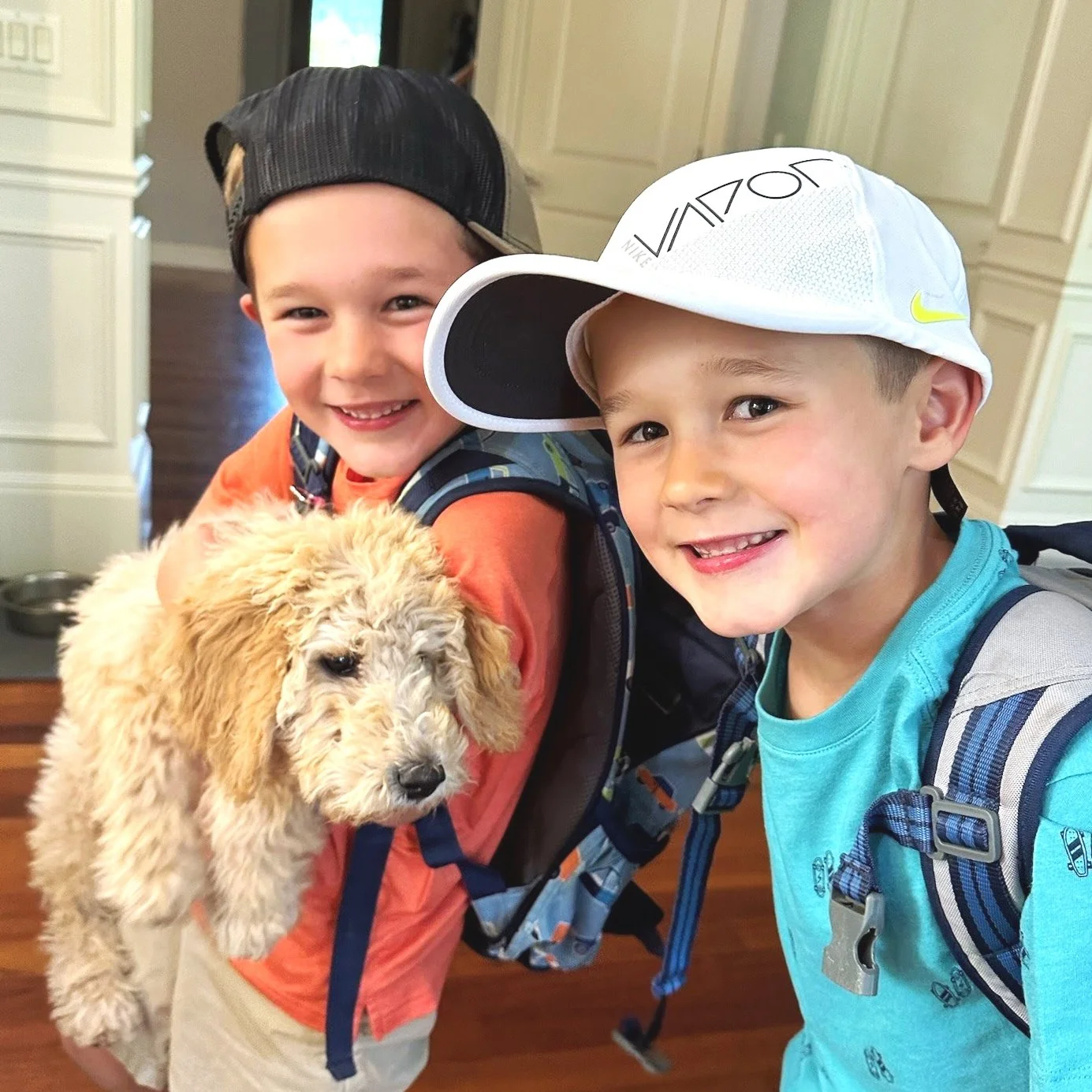 Two smiling young boys with backpacks, one wearing a black cap backwards, the other a white cap, holding a fluffy golden doodle puppy, indoors with cream-colored walls and wooden flooring.