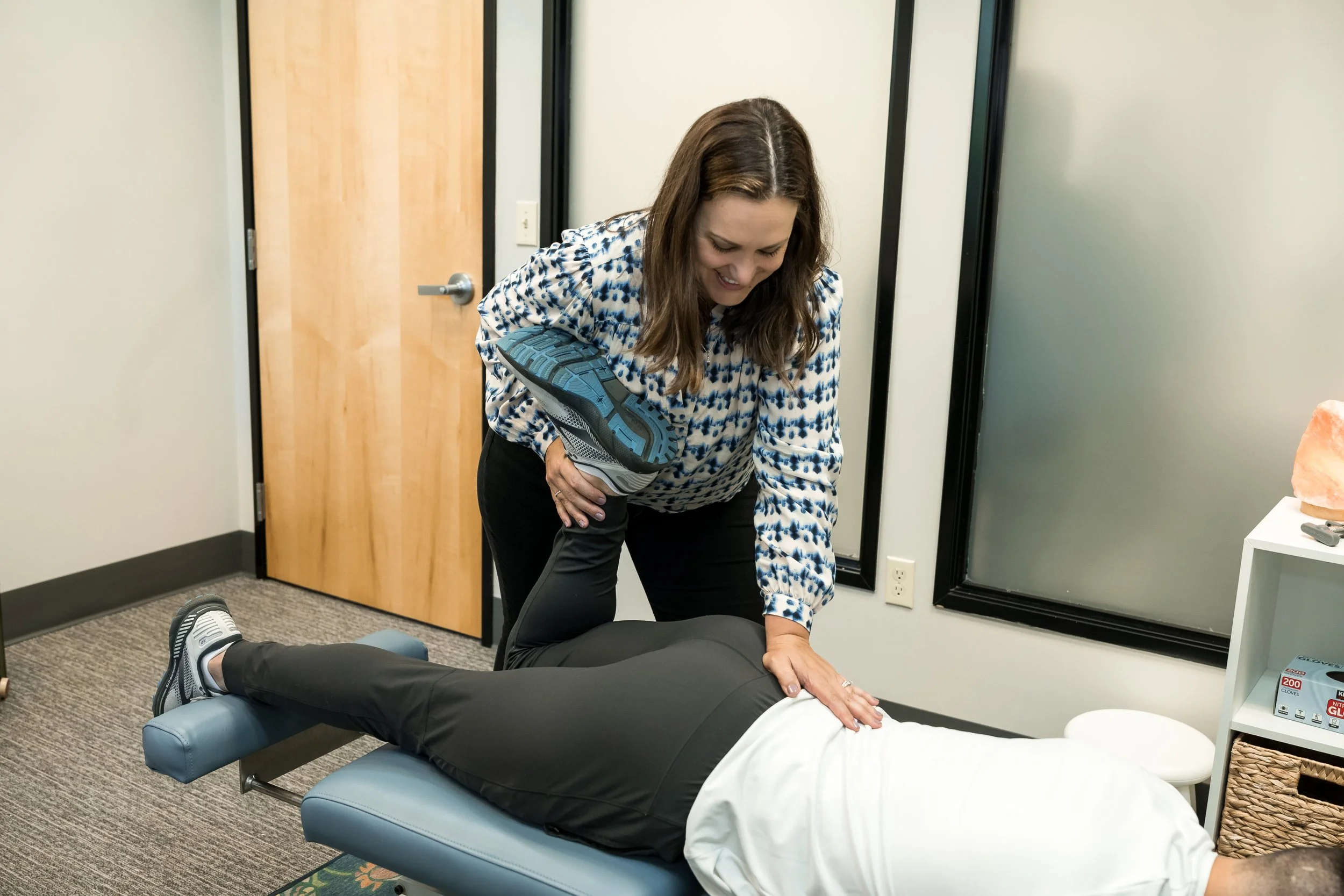 A woman is performing a physical therapy exercise on a person lying face down on a therapy table, holding a shoe and smiling. The room has a closed wooden door, a window, and medical supplies.