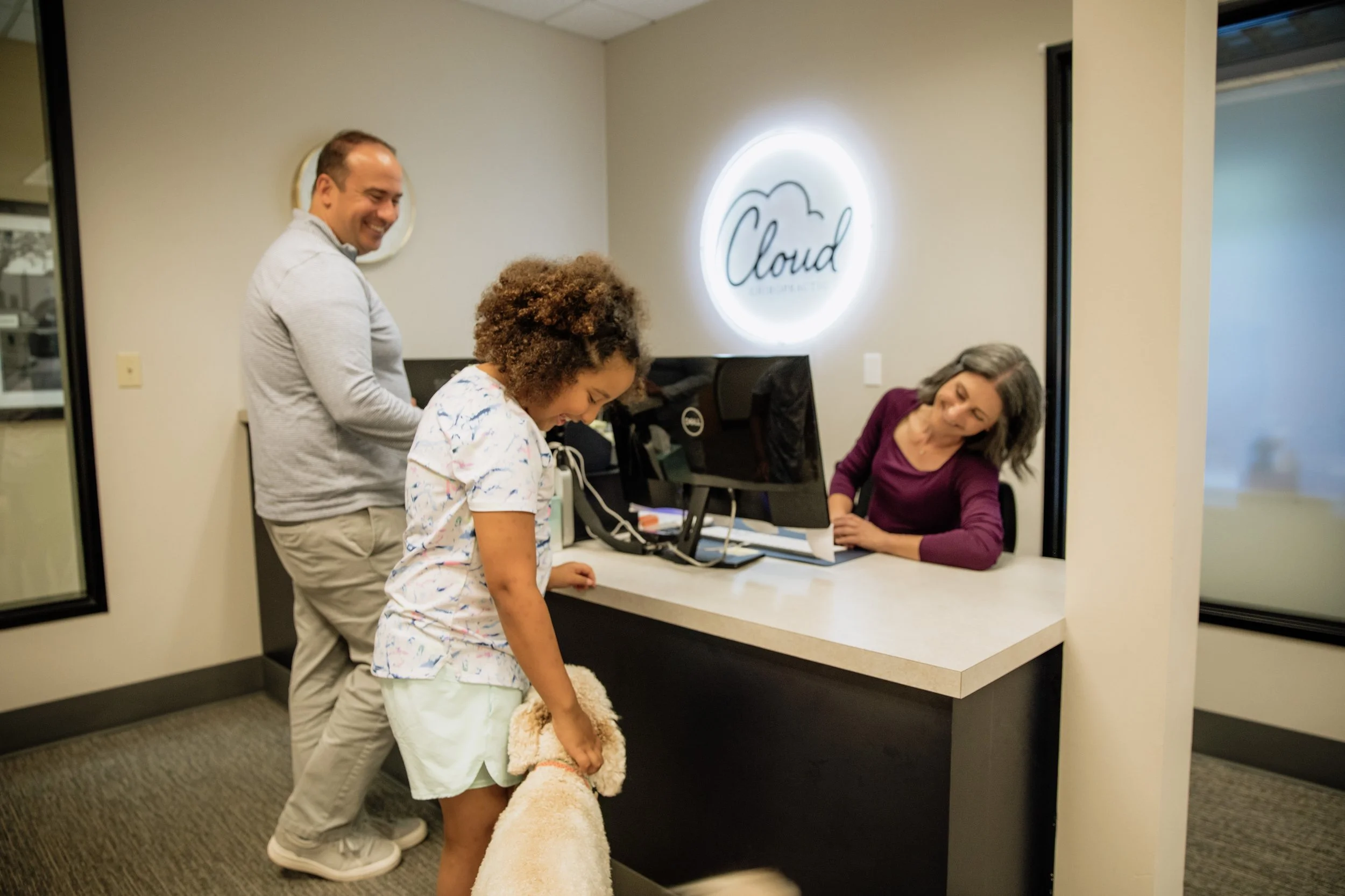 Three people in a healthcare setting, a man, a woman, and a young girl with a teddy bear, at a reception desk with a sign that reads 'Cloud' on the wall behind them.