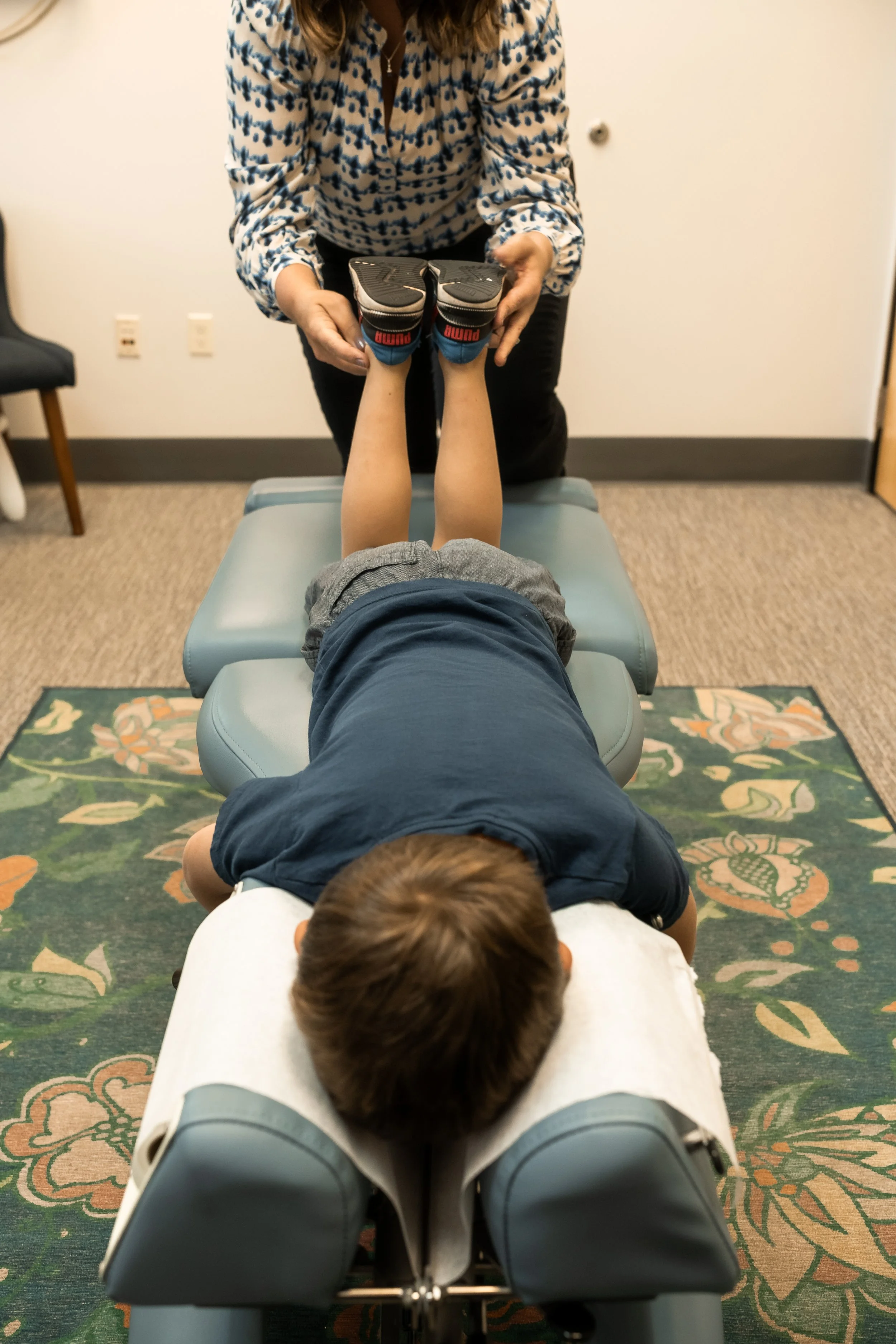 Child lying face down on a chiropractic adjustment table, receiving treatment from a female practitioner in a clinical office setting.