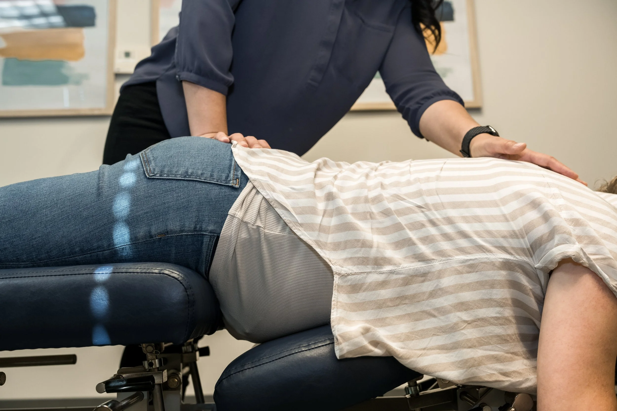 A person lying face down on a medical examination table while a healthcare professional performs a chiropractic adjustment or massage. The person is wearing jeans and a striped shirt, and the healthcare professional is dressed in dark clothing and a 