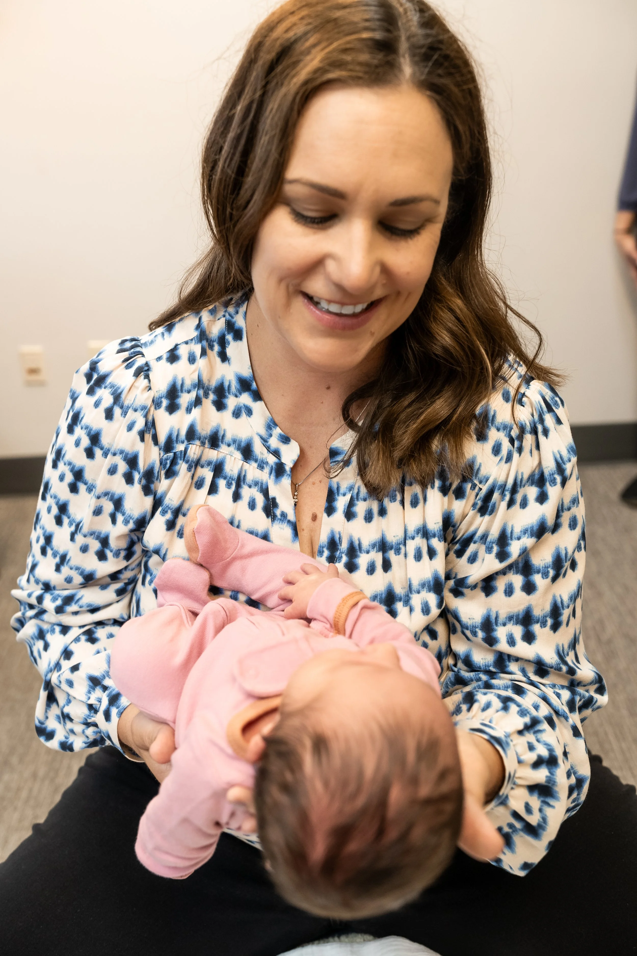 A woman with brown hair smiling while holding a baby in pink clothing.