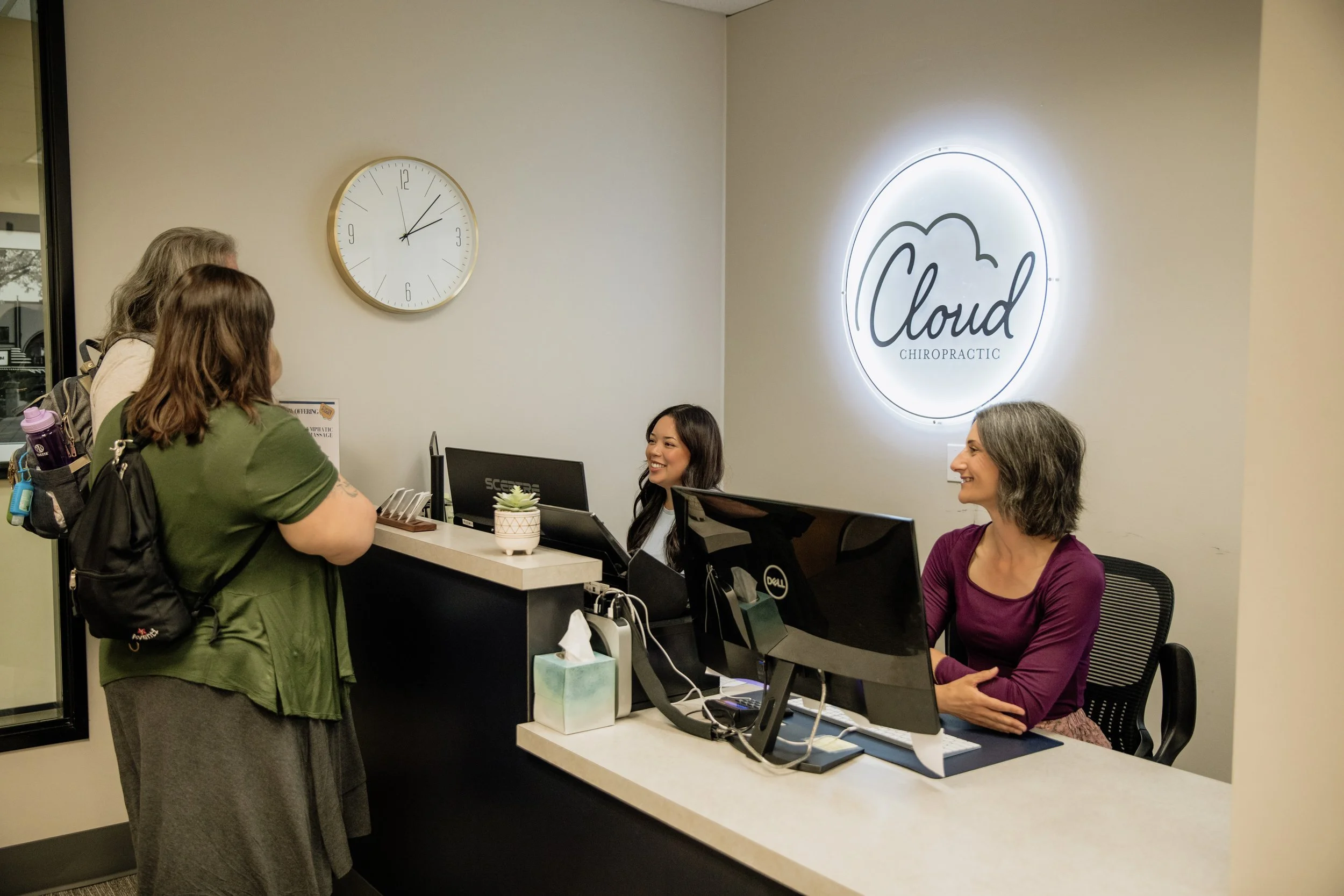 A woman and a girl check in at a chiropractic office reception desk, engaging in conversation with two staff members seated behind the counter. The office has a large illuminated sign reading 'Cloud Chiropractic' and a wall clock showing the time as 