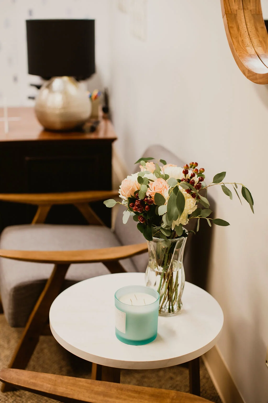 A bouquet of pink roses, white flowers, eucalyptus leaves, and red berries in a glass vase on a small white table. A light blue candle in a glass holder is next to it. The background shows a wooden chair with a gray cushion, a wooden desk, a beige la