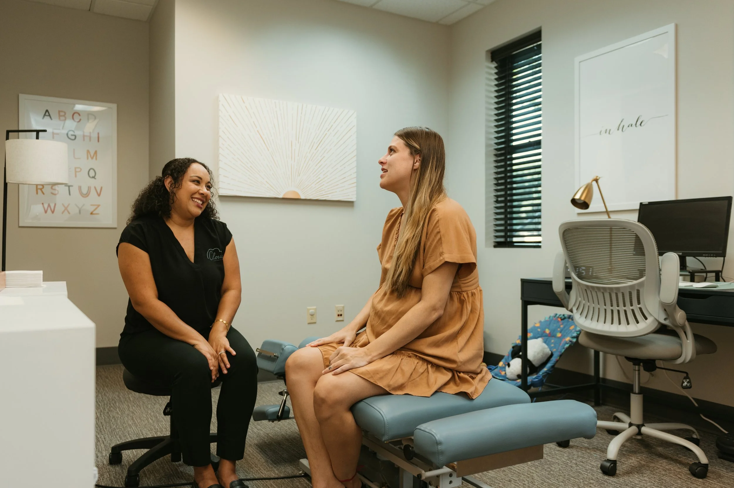 A healthcare professional and a woman sitting on an exam table in a consultation room, engaging in conversation. The room has medical posters and a laptop on a desk.