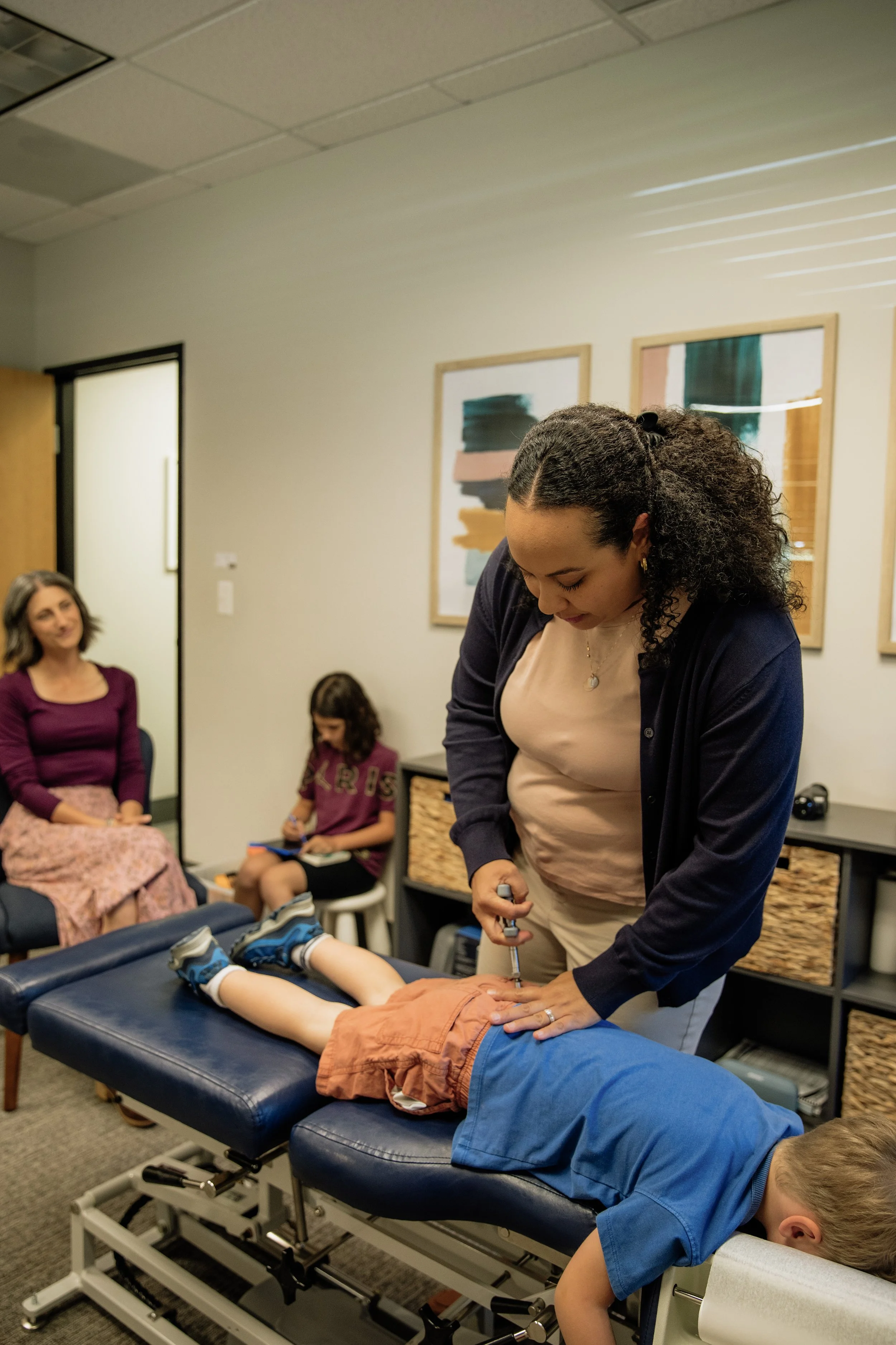A chiropractor or physical therapist administers a treatment on a young boy lying face down on a chiropractic table, while other children and a woman are seated in a room decorated with colorful abstract paintings.