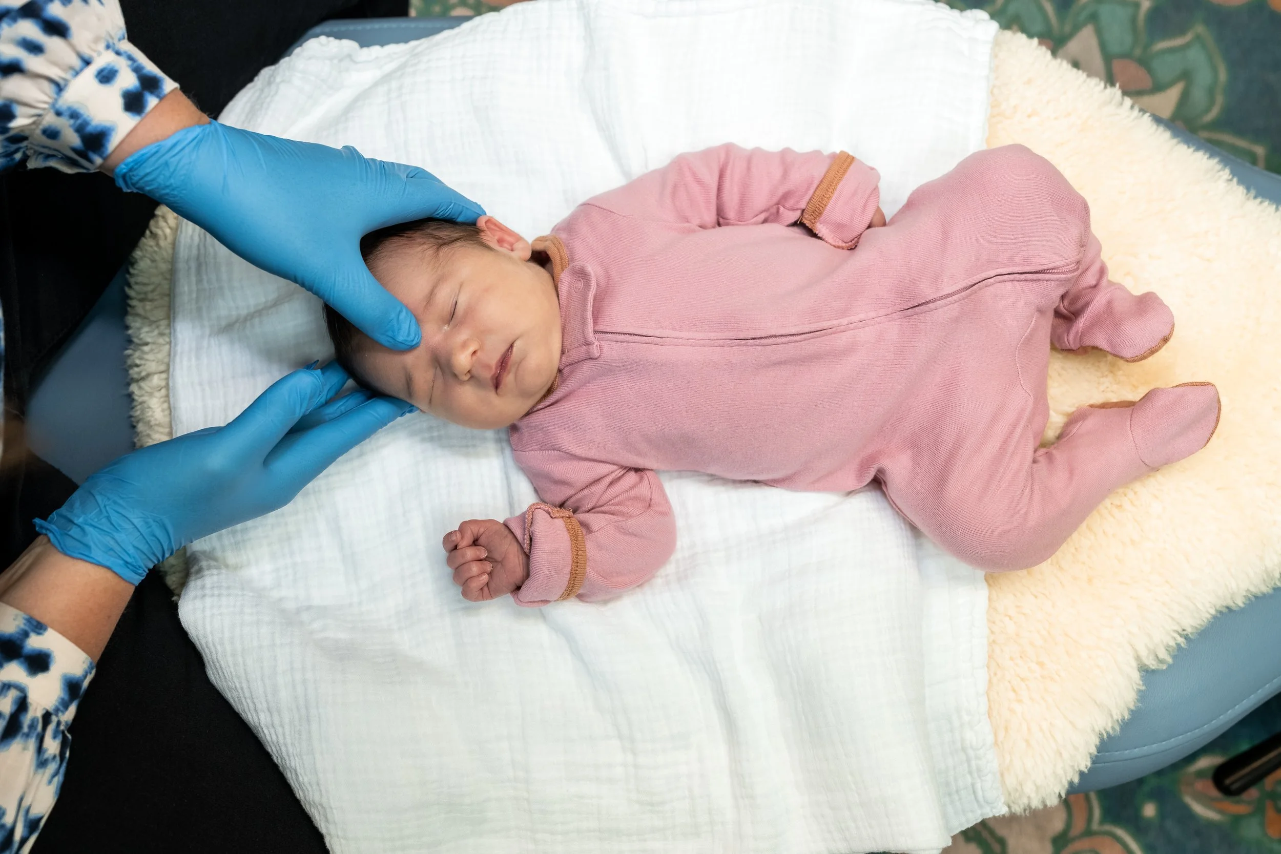 A sleeping baby lying on a changing table with a white sheet and a soft yellow blanket, with someone in a blue glove gently touching the baby's head.