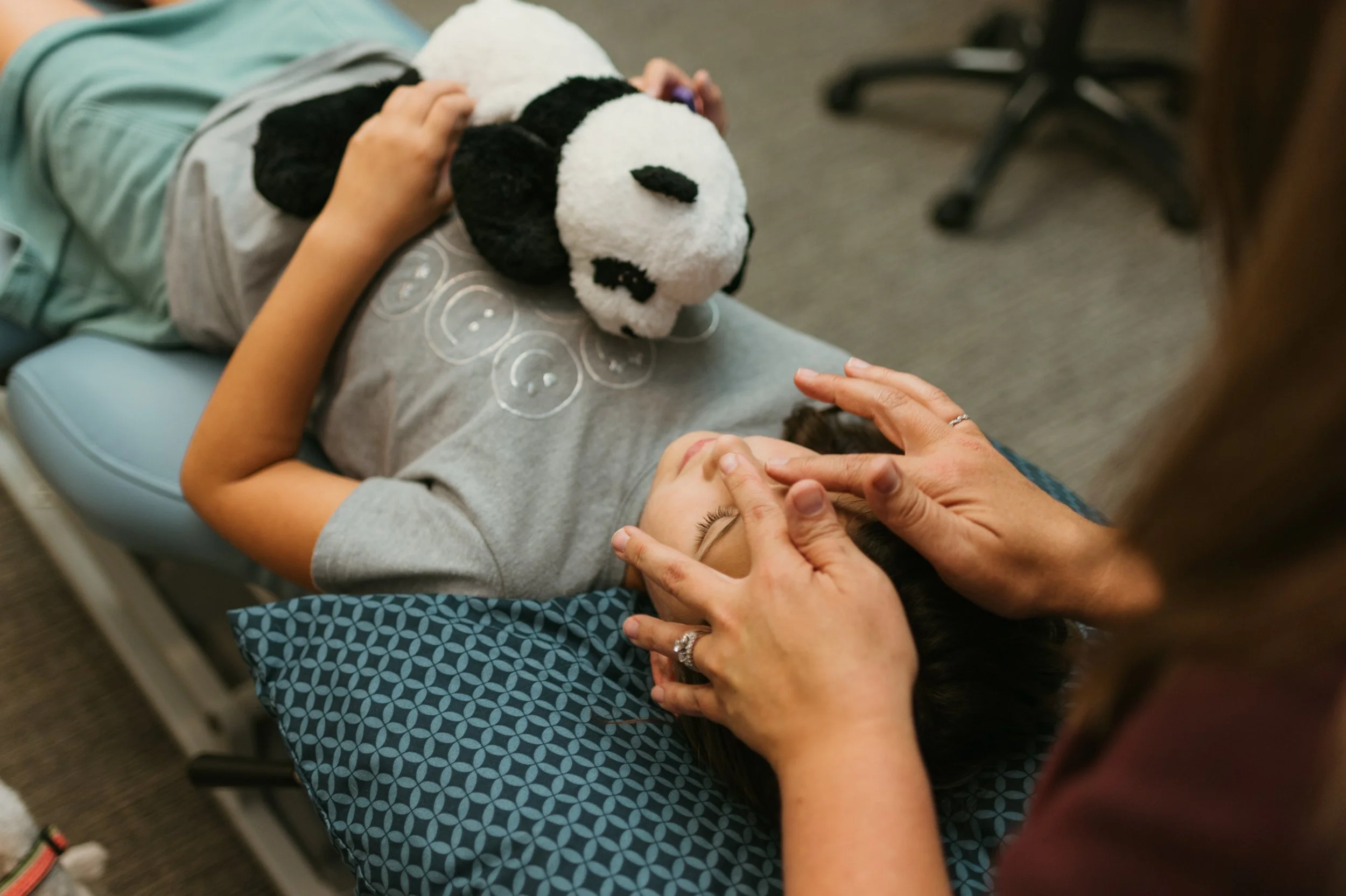 A child is lying on a bed, holding a stuffed panda bear on their chest, while an adult, likely a healthcare provider, gently touches their forehead and face, possibly during a check-up or comforting moment.