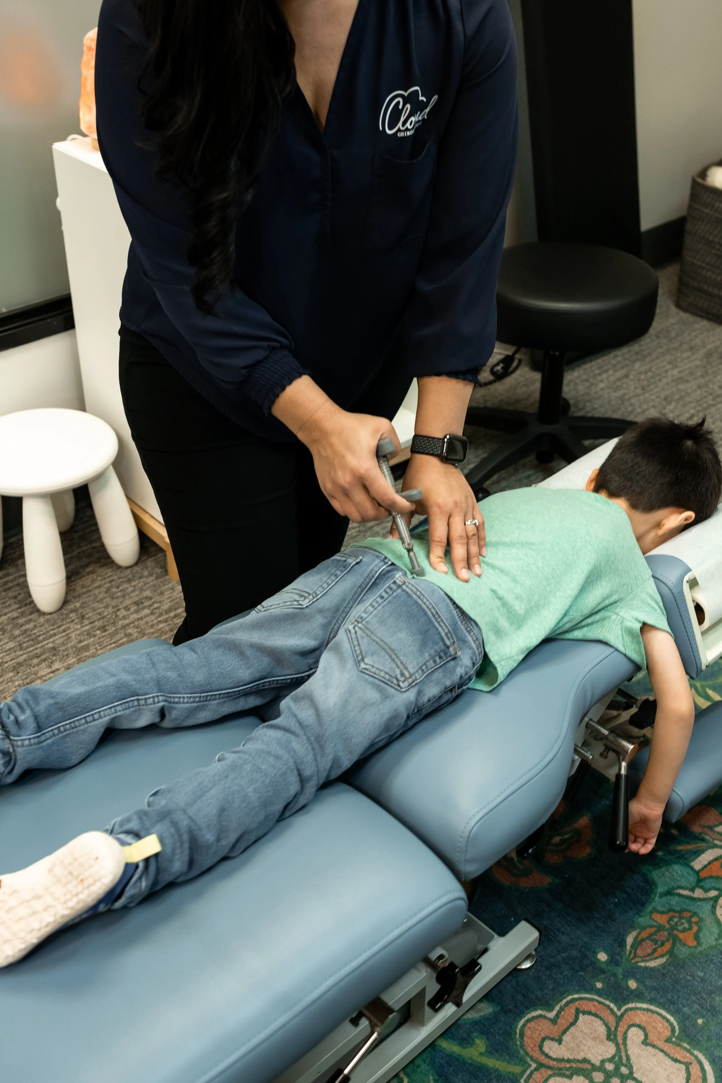 A person lying face down on a medical examination table receiving an injection in the lower back or hip area from a healthcare professional. The healthcare worker is wearing a navy shirt with a logo on the chest and a black watch on their wrist.