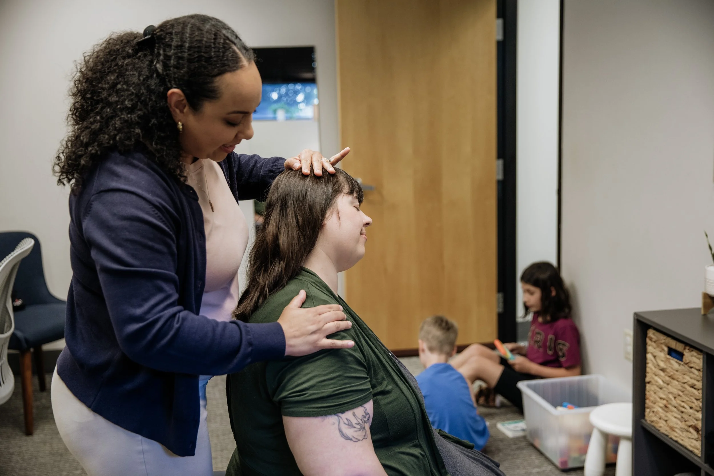 A woman with curly hair gently places her hand on another woman's head, who is seated with eyes closed in a therapeutic or healing setting. In the background, children are sitting and engaged in activities.