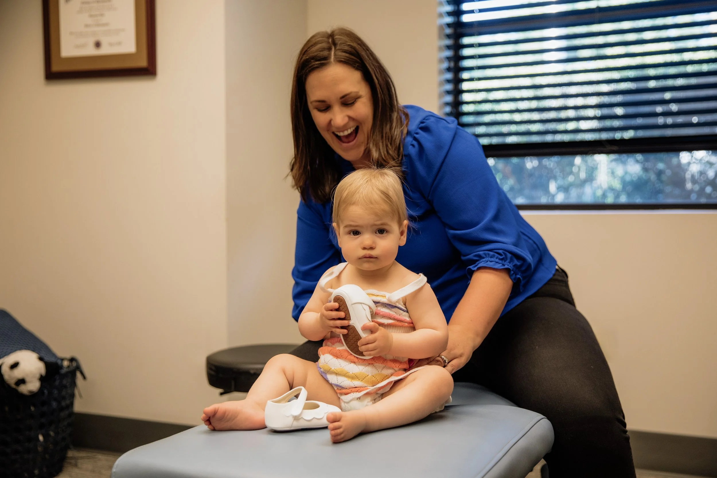 A woman with brown hair wearing a blue shirt is smiling while holding a young girl on her lap in a room with a window and framed certificate on the wall. The girl has blonde hair, wears a colorful striped dress, and is holding a small shoe.