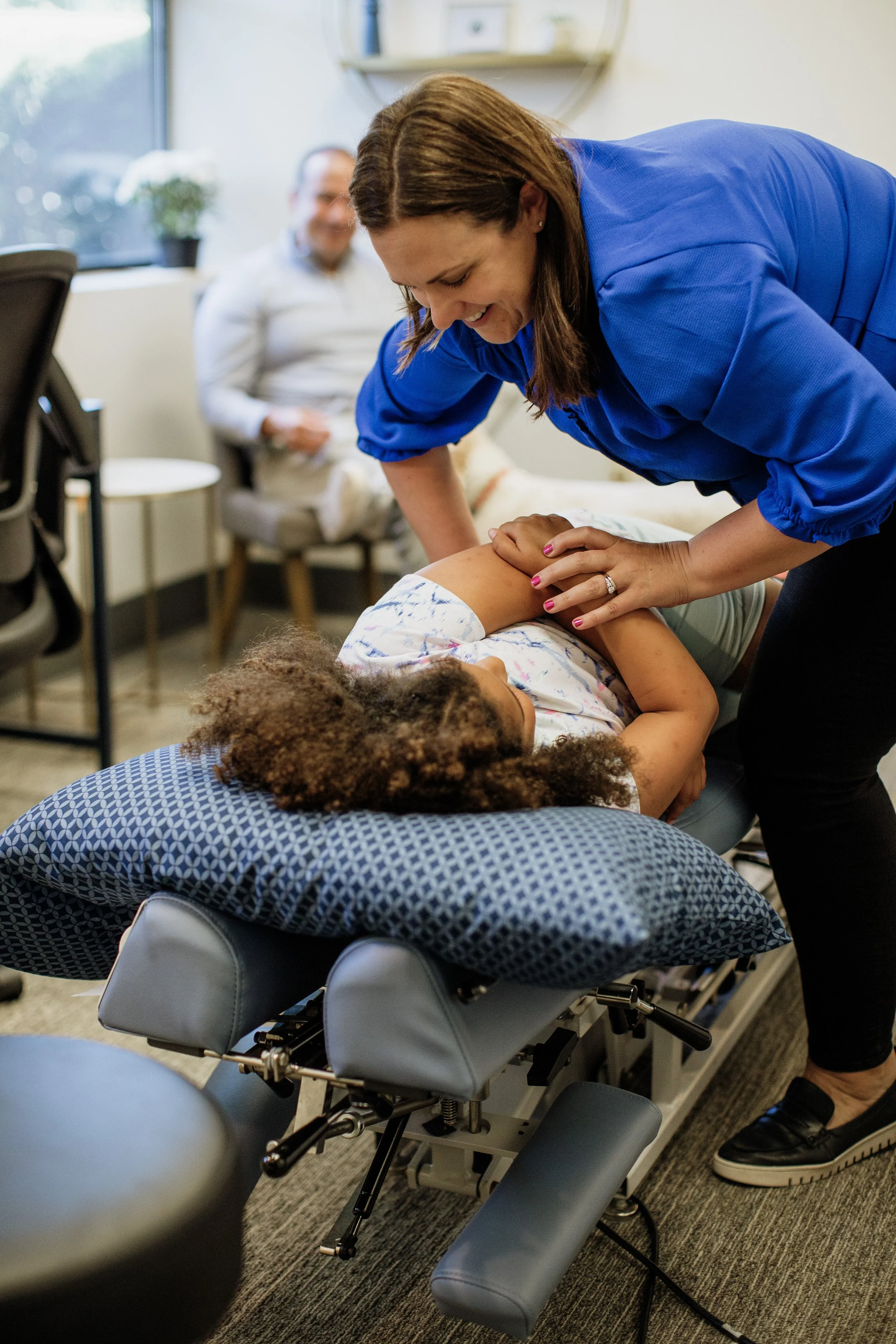 A woman receiving chiropractic adjustment from a healthcare professional in an office setting, with a person sitting in a chair and a window in the background.