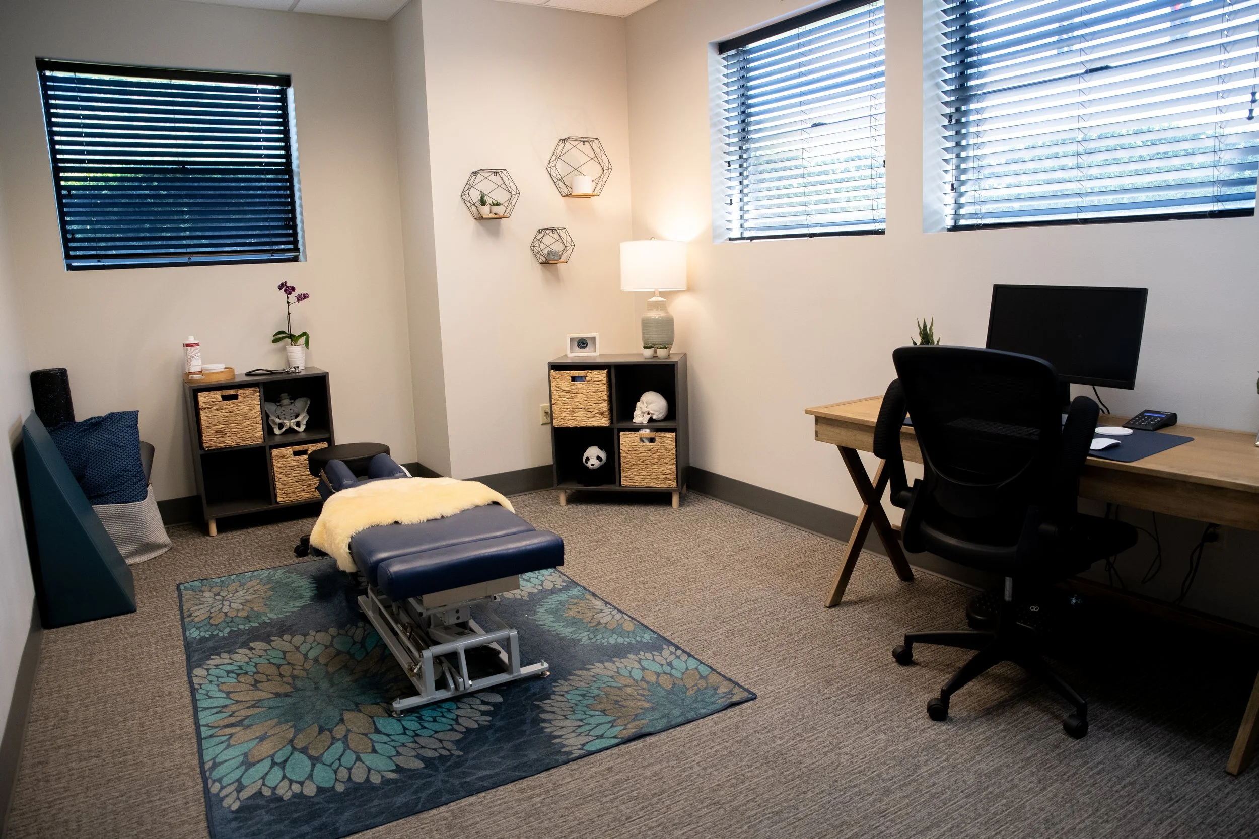 A therapy or massage treatment room with a chiropractic adjusting table, a computer on a wooden desk, and various decorative items on shelves, including plants, a skull model, and a panda plush.