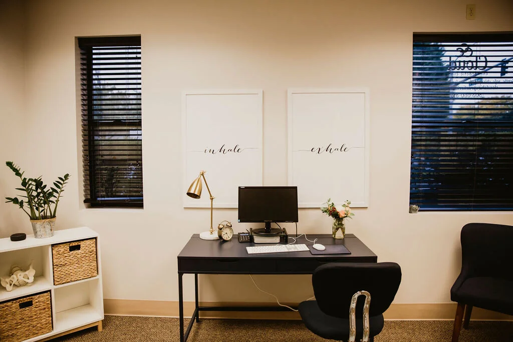 Home office with a black desk, desktop computer, lamp, clock, flower vase, and window blinds.