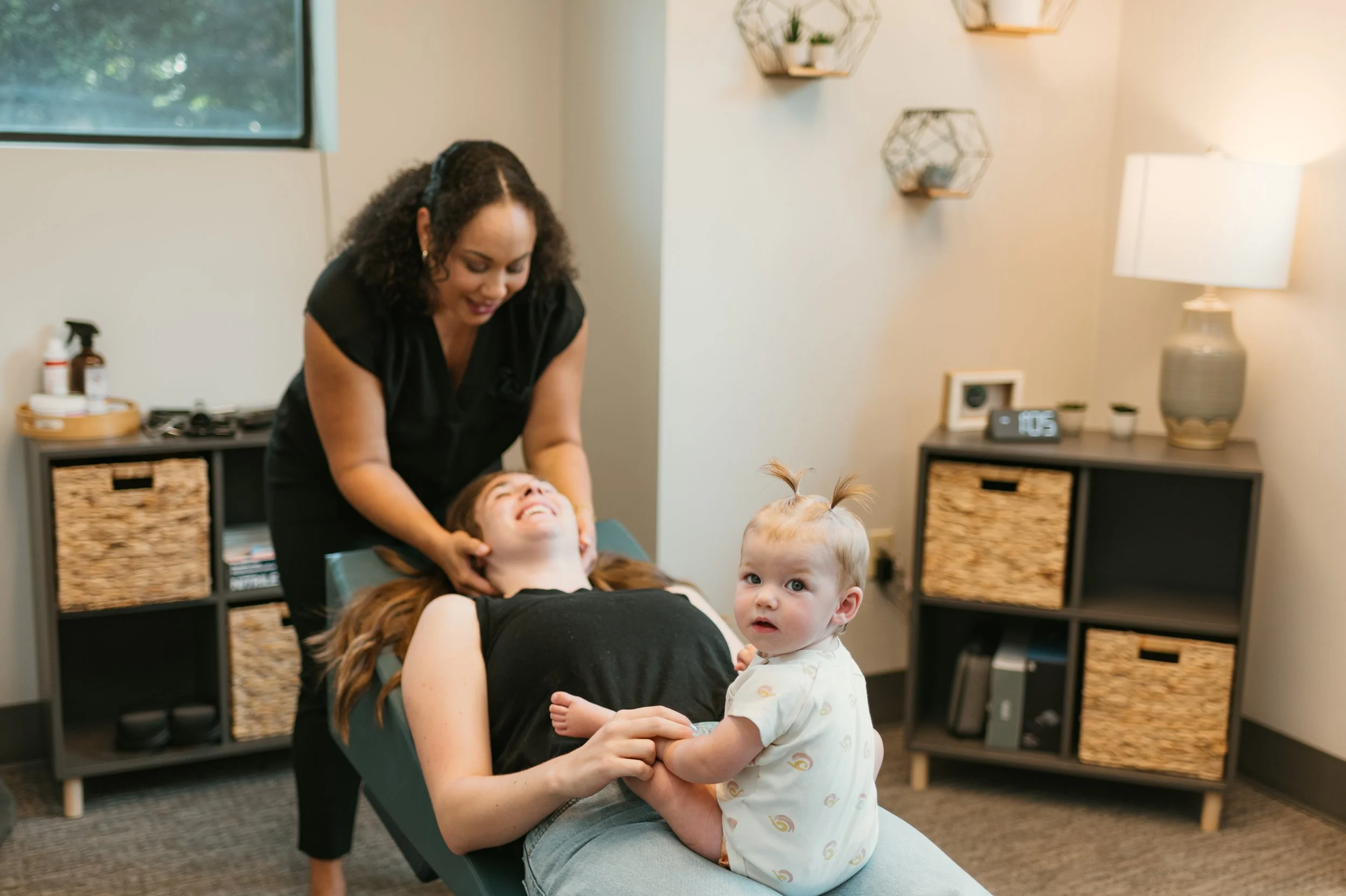 A woman receiving a massage while lying on a massage table, with a young child standing next to her holding her hand, and another child with pigtails looking at the camera. The room has a window, wall-mounted shelves with plants, a bedside table with