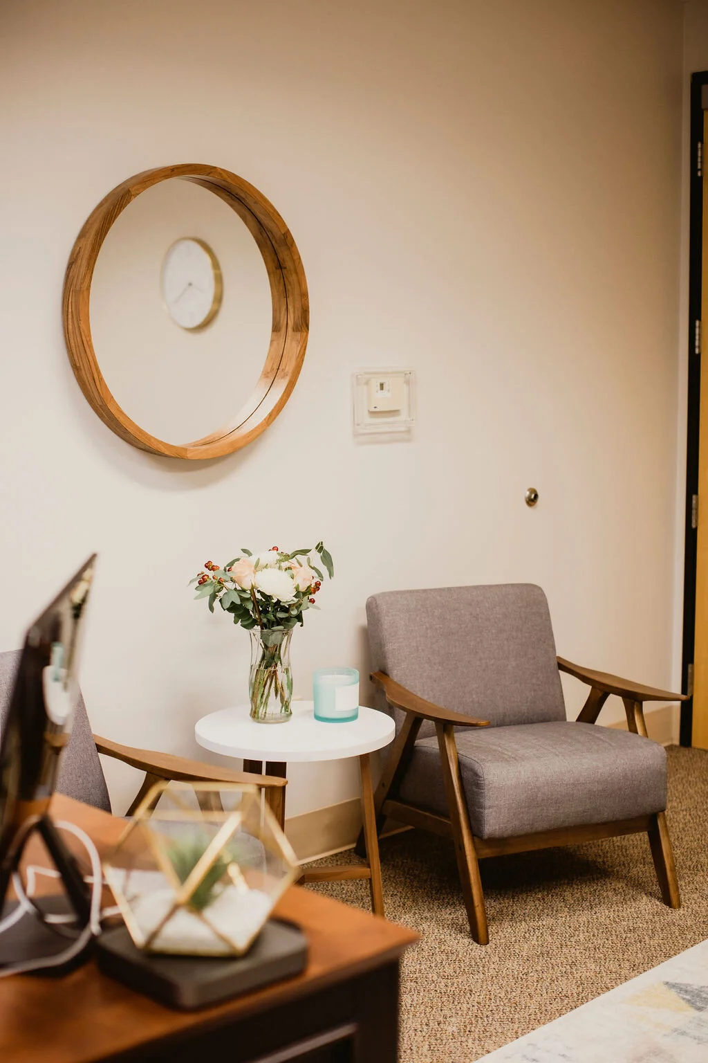 A cozy waiting room with a gray armchair, a white side table with a flower vase and a candle, a circular wooden mirror on the wall, and a wall clock reflected in the mirror.