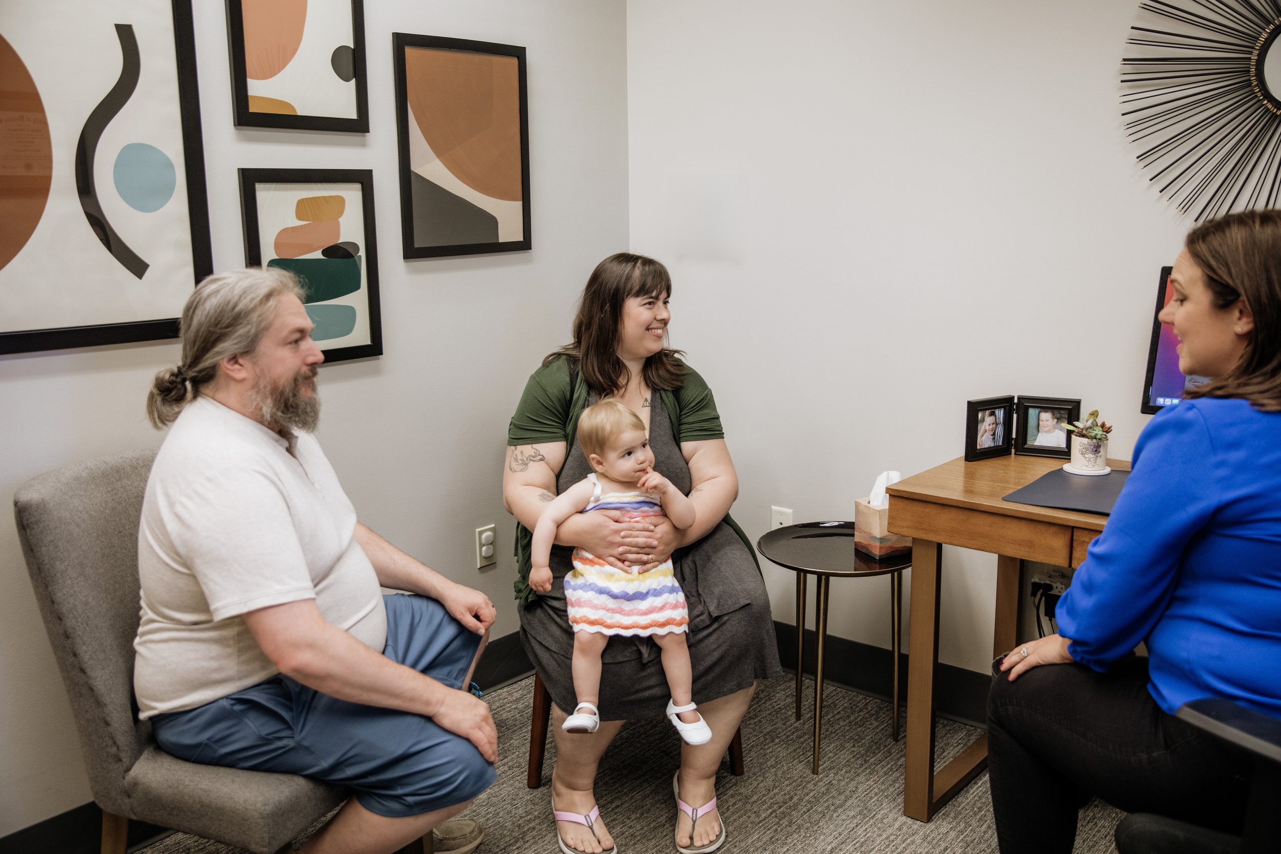 A group of four people, including a man with gray hair and a beard, a woman holding a child, another woman with brown hair, and an unseen person, are seated and standing in an office or waiting room. The woman with the child is smiling and the child 