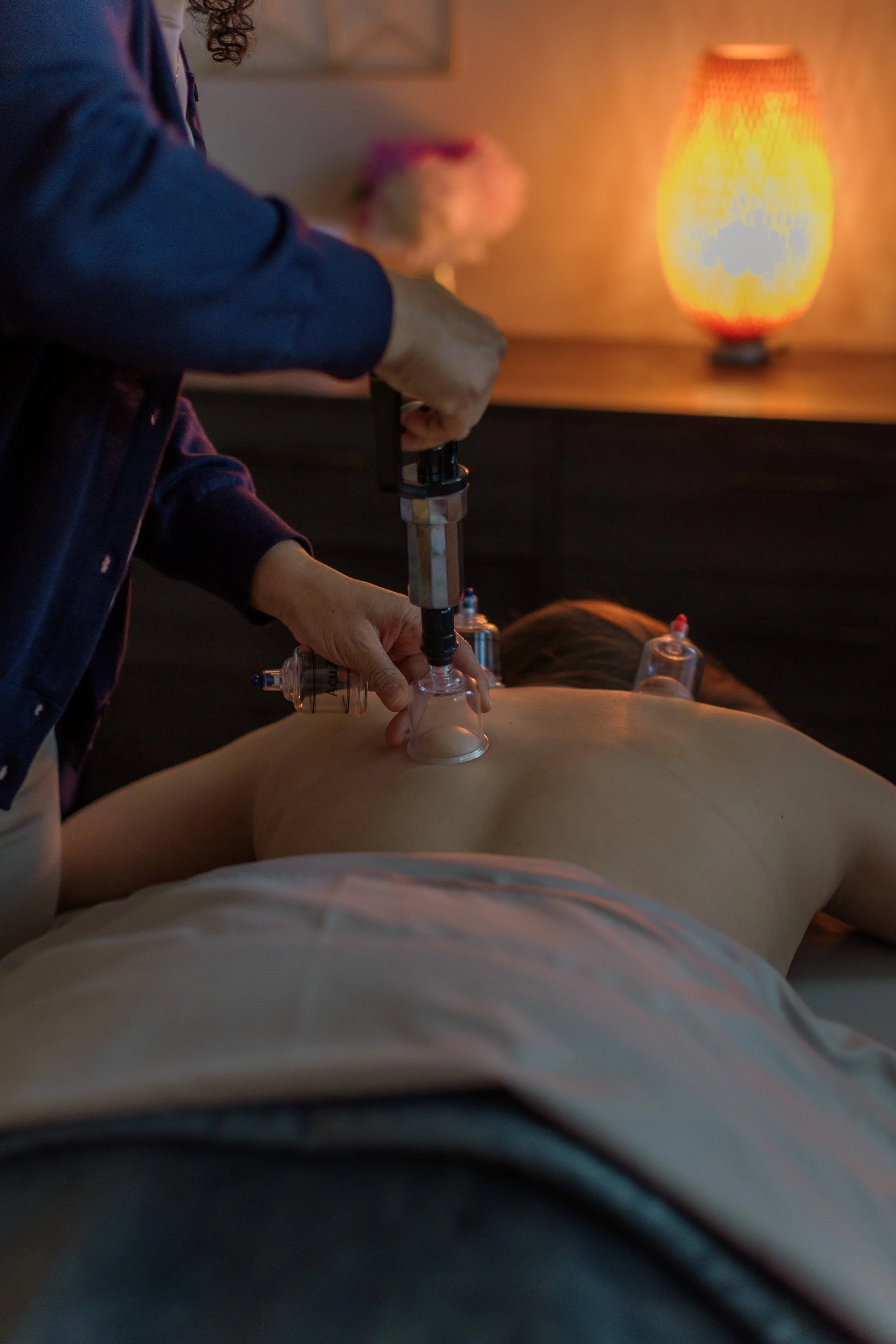 A person lying face down on a massage table receiving cupping therapy, with glass cups placed on their back. The scene is softly lit with a warm orange lamp in the background.