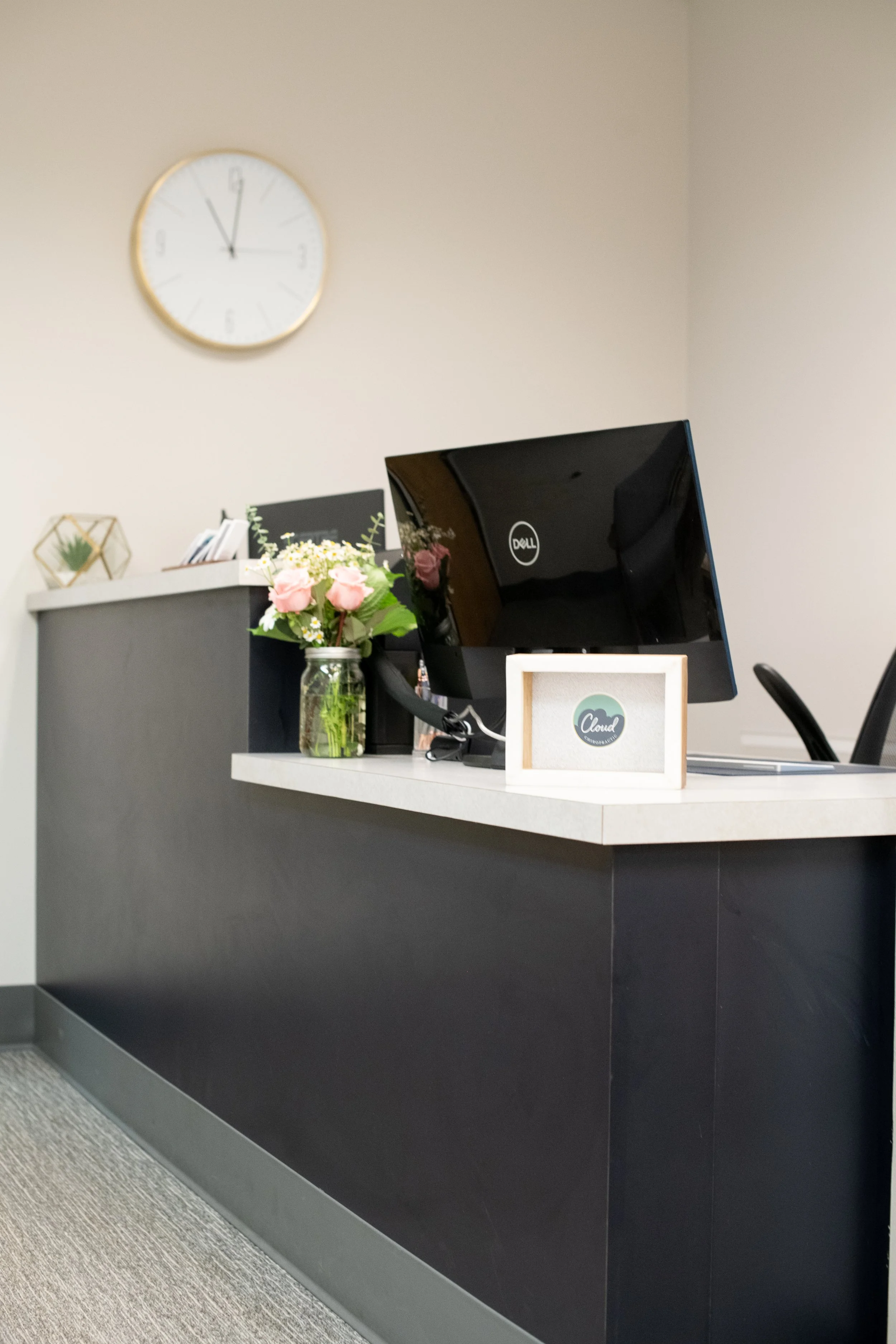 Office reception area with a black desk, a computer monitor, and a small framed sign that reads 'Cloud.' There is a glass jar of pink and white flowers on the desk. A white wall clock shows the time as 11:58. The wall behind the desk is beige, and th