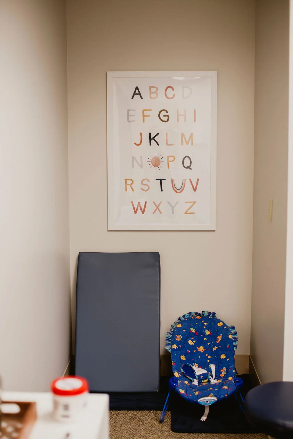 A small medical examination room with a beige wall, a framed alphabet chart, a black padded examination table, and a blue baby bouncer with colorful fish patterns.