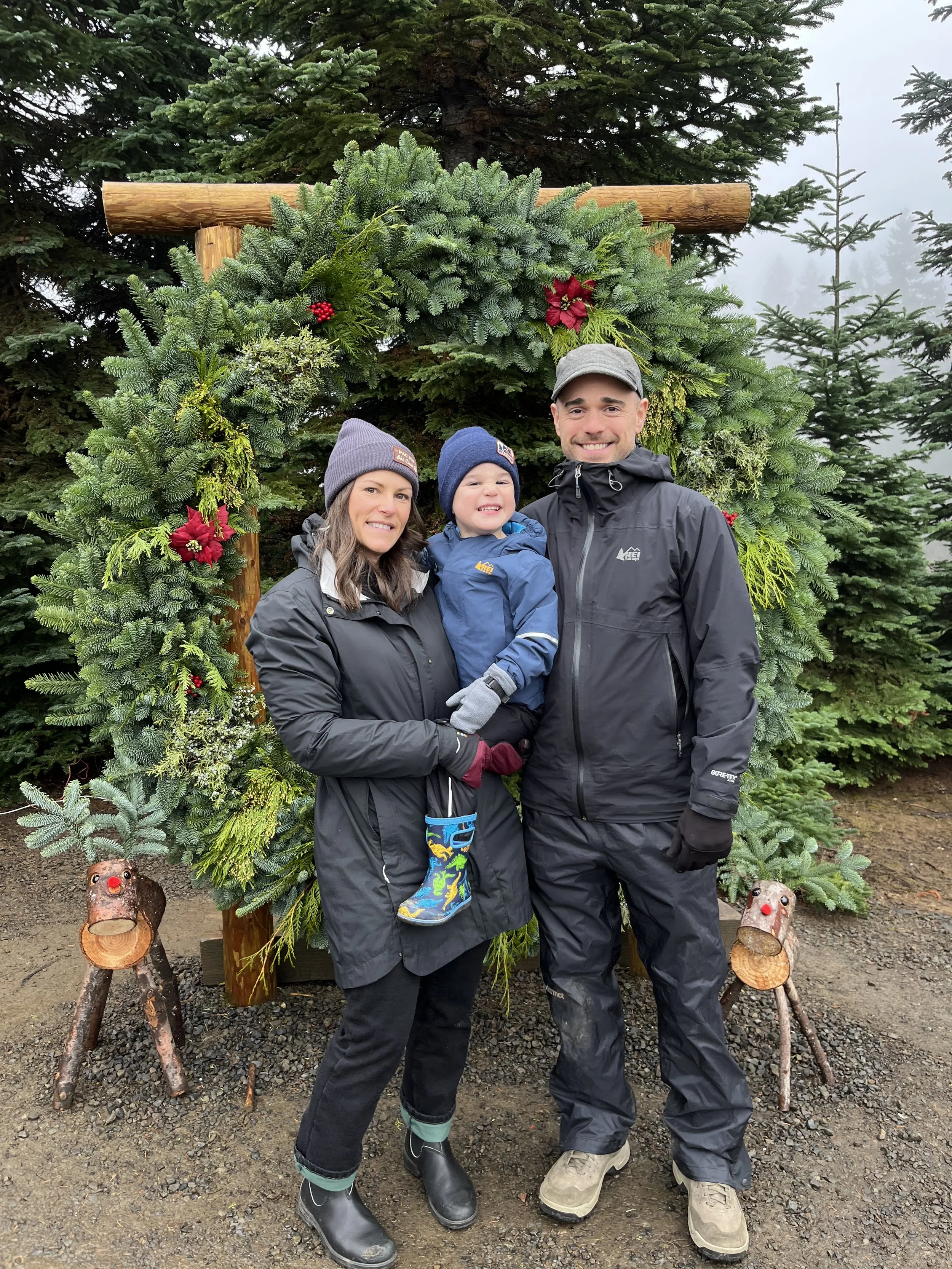 A family of three stands in front of a decorated Christmas tree outdoors. The woman has brown hair, wears a black jacket and gray beanie. The boy has blonde hair, wears a blue jacket and beanie, and is smiling. The man has short hair, wears a black jacket and gray cap. There are two small wooden reindeer figures in front, and the scene is foggy with evergreen trees in the background.
