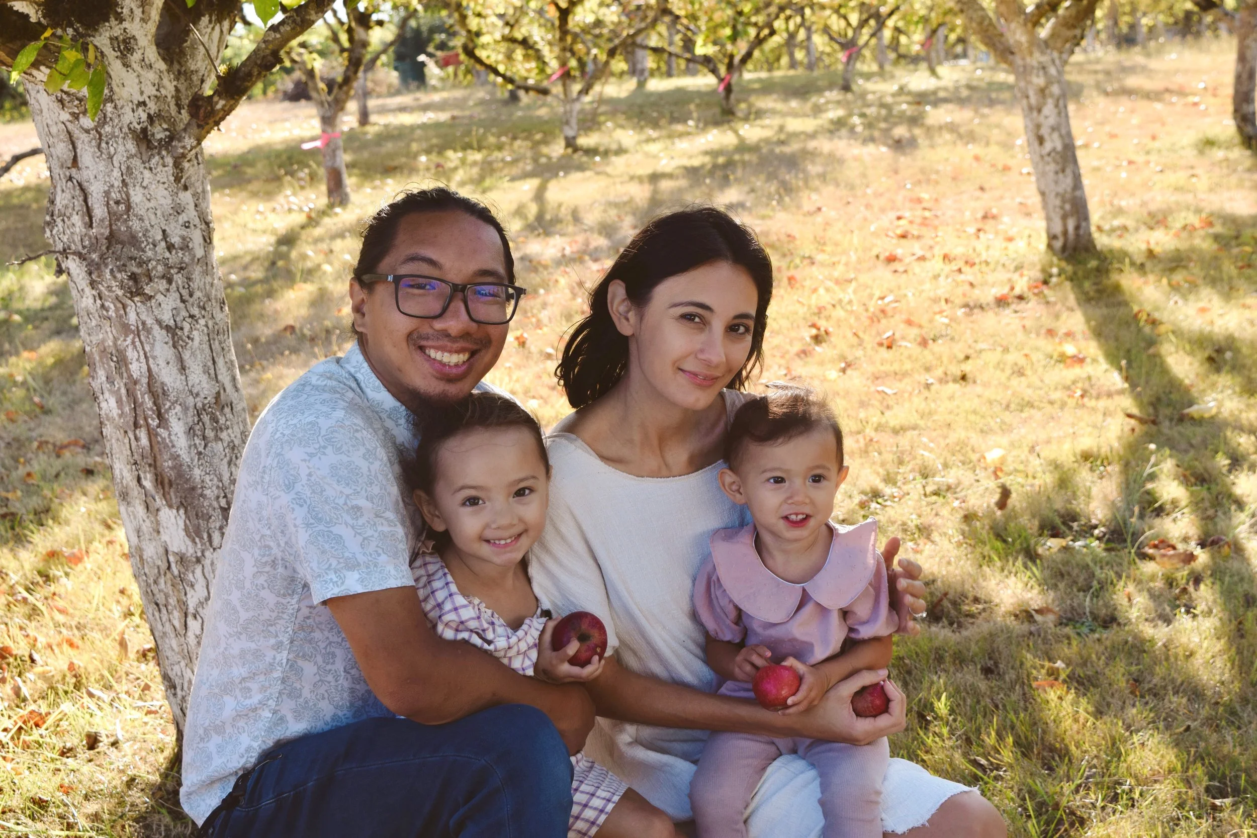 A family of five sitting outdoors under a tree in an orchard during fall, holding apples and smiling.