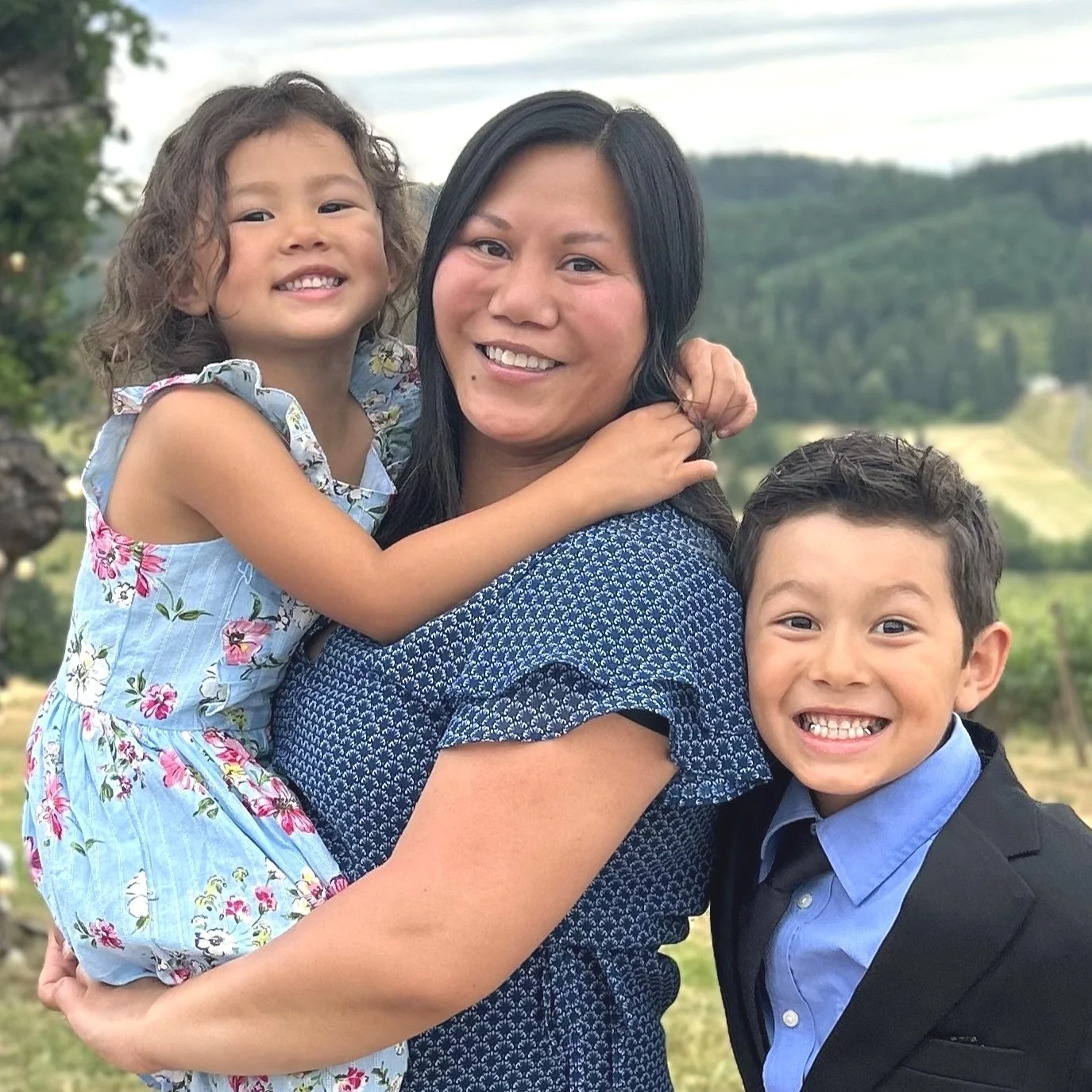 A woman holding a young girl and a young boy outside with a blurred green landscape in the background. The girl is wearing a blue floral dress and smiling, while the boy is dressed in a blue shirt and blazer, also smiling.