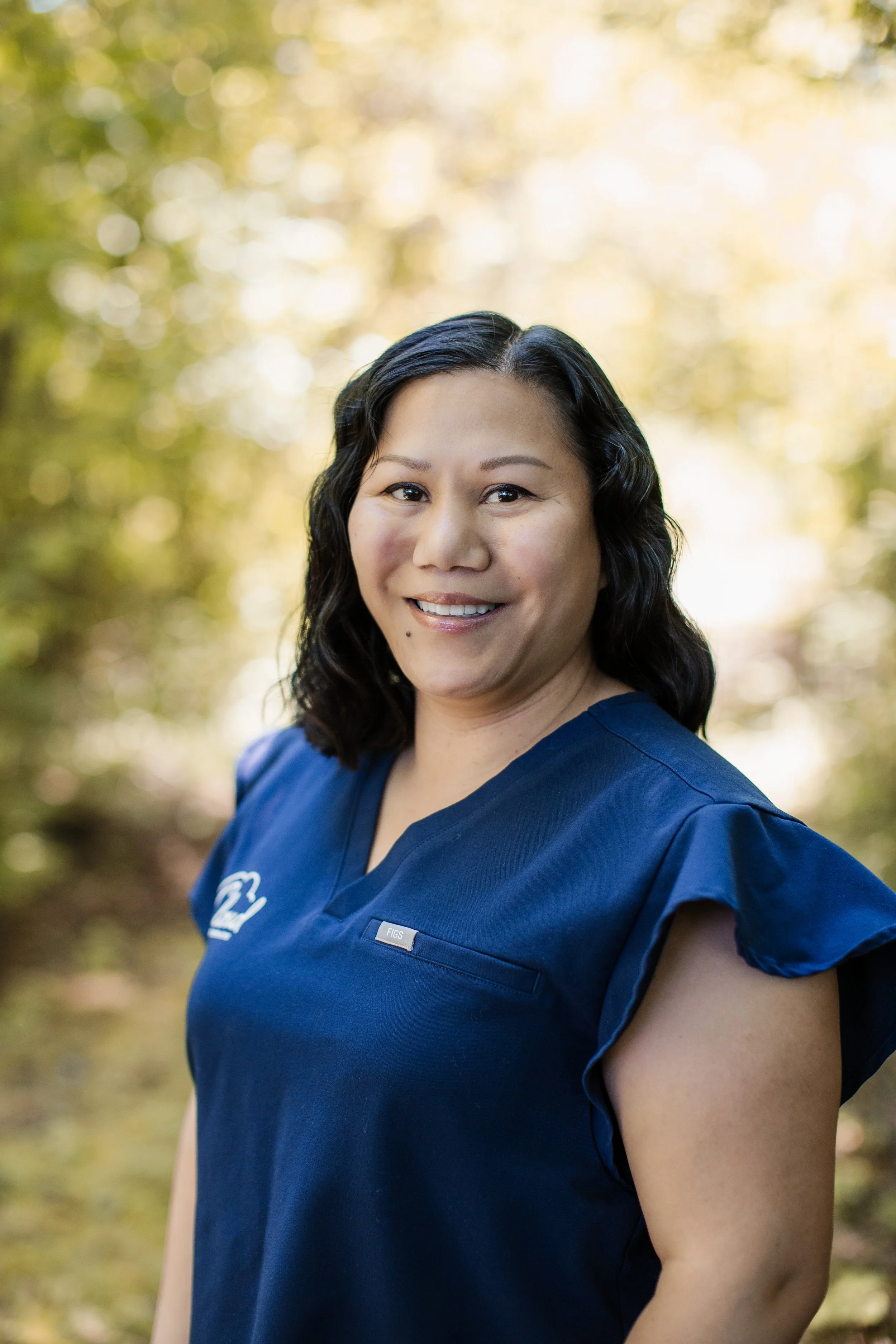 A woman in a blue medical uniform smiling outdoors, with a blurred background of green and yellow foliage.
